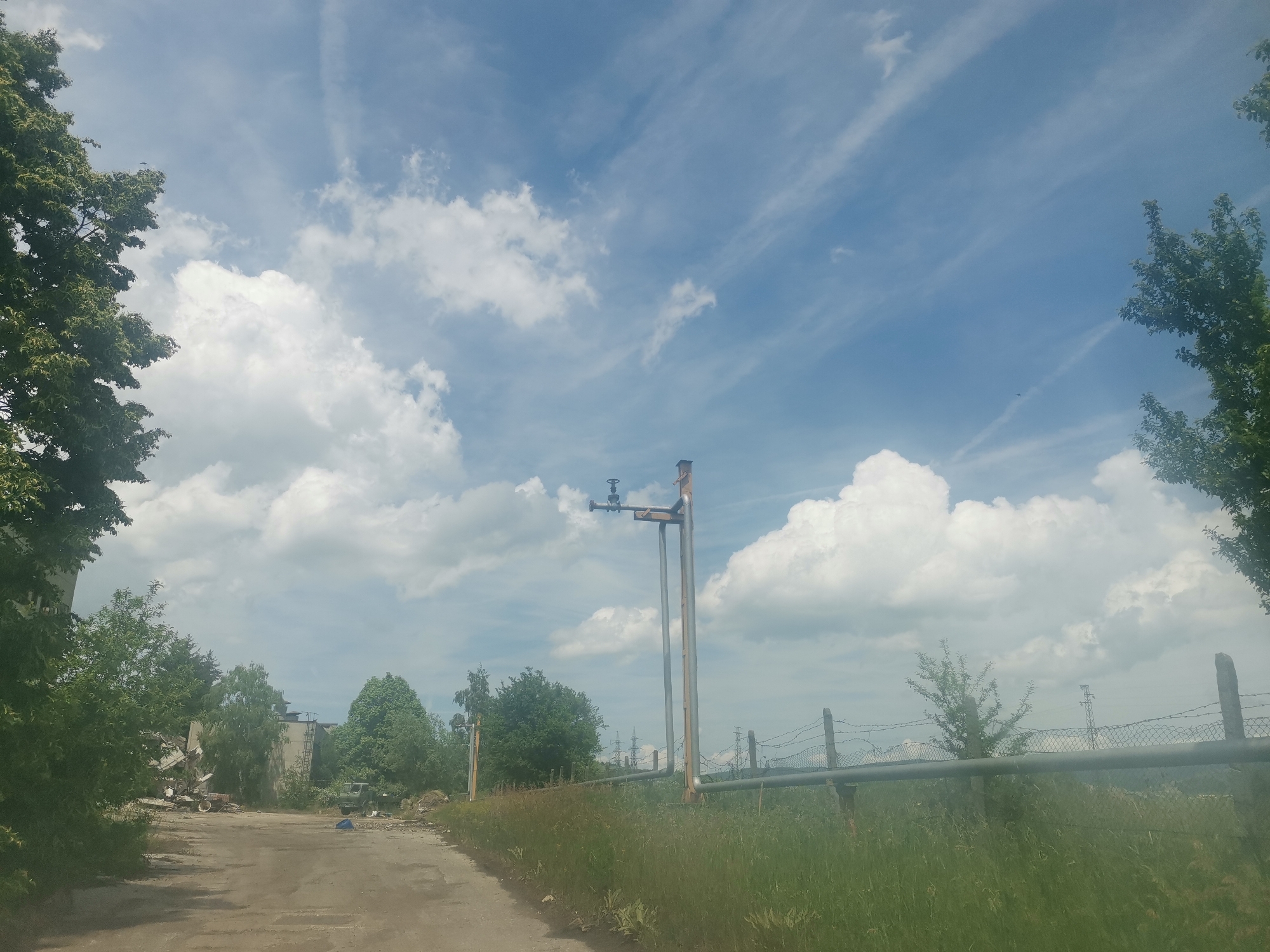 The image captures a sunny daytime scene in Zhivkovo, Bulgaria, under a bright blue sky with scattered white cumulus and wispy cirrus clouds. A worn, unpaved road, likely a dirt or gravel track, runs along the left side of the frame, flanked by green grass and weeds.
On the left, beyond dense green trees and bushes, lie dilapidated or partially collapsed structures, possibly industrial or agricultural buildings. A white vehicle, resembling a tractor or similar equipment, is visible near these ruined structures, with a green truck also discernible further back among the trees.
To the right, a prominent section of elevated utility piping, supported by metal posts, stretches across the midground. A valve mechanism is visible on the pipe. Below this pipeline, a chain-link fence with concrete posts and barbed wire runs horizontally, bordered by tall, wild green grass. Faint power line towers are visible in the distant background on the right, interspersed with trees.
No people are visible in the image, and there is no discernible text. The scene suggests a neglected or abandoned rural or semi-industrial area, evoking a sense of decay amidst the natural growth.