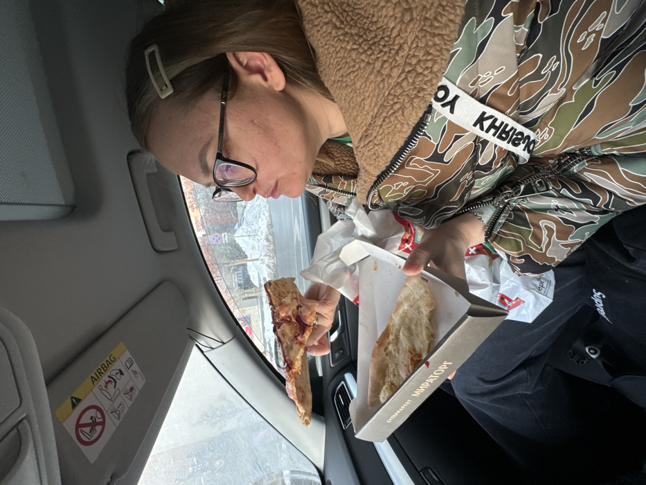 A female individual is depicted seated in the front passenger seat of a vehicle, eating pizza. She wears black-framed glasses, a light brown hair clip on her right side, and a multi-colored camouflage-patterned jacket with a brown fleece collar. Her right hand holds a large slice of pizza with a reddish topping. Her left hand supports an open rectangular cardboard pizza box, labeled "СУПЕРМАРКЕТ МИРАТОРГ" (SUPERMARKET MIRATORG), which contains a second, plainer pizza slice. A white plastic bag with red markings, likely from Miratorg, is partially visible beneath the box. The car's interior features a gray headliner and a sun visor with an "AIRBAG" warning label. Through the vehicle's windows, a winter outdoor scene is visible, characterized by snow accumulation on the ground, bare trees, and distant buildings. Road signs are partially discernible in the background. The presence of the "Мираторг" supermarket branding indicates a Russian location, consistent with the city of Moscow.