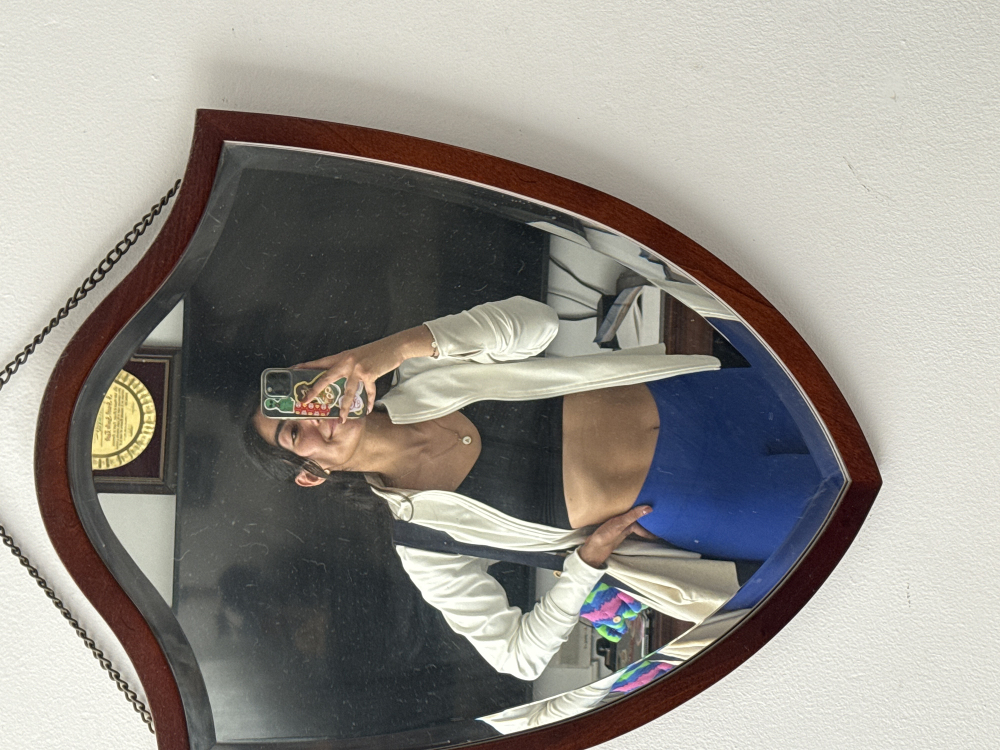 A young woman is captured in a mirror selfie, reflecting in a distinctive leaf-shaped mirror with a dark wooden frame and a hanging metal chain, set against a white, textured wall. She holds her phone, which has a colorful, patterned case, to take the picture. The woman has dark hair, a slight, relaxed smile, and is looking directly at her phone's camera.
She is wearing a light-colored, possibly off-white, long-sleeved top or jacket over a dark top or bra, with blue bottoms. A delicate necklace with a small, light-colored pendant adorns her neck, and a subtle bracelet is visible on her right wrist.
The reflection within the mirror shows glimpses of the surrounding indoor environment. To the left, behind her head, a framed, circular item resembling a medal or plaque is partially visible. In the lower right corner of the mirror's reflection, colorful patterned fabric can be seen. The mirror surface appears slightly dusty. The scene depicts a casual, everyday moment in an indoor setting, likely in Puebla, Mexico, as provided by the location context. No clear text is legible on the phone case or the framed item in the reflection.
