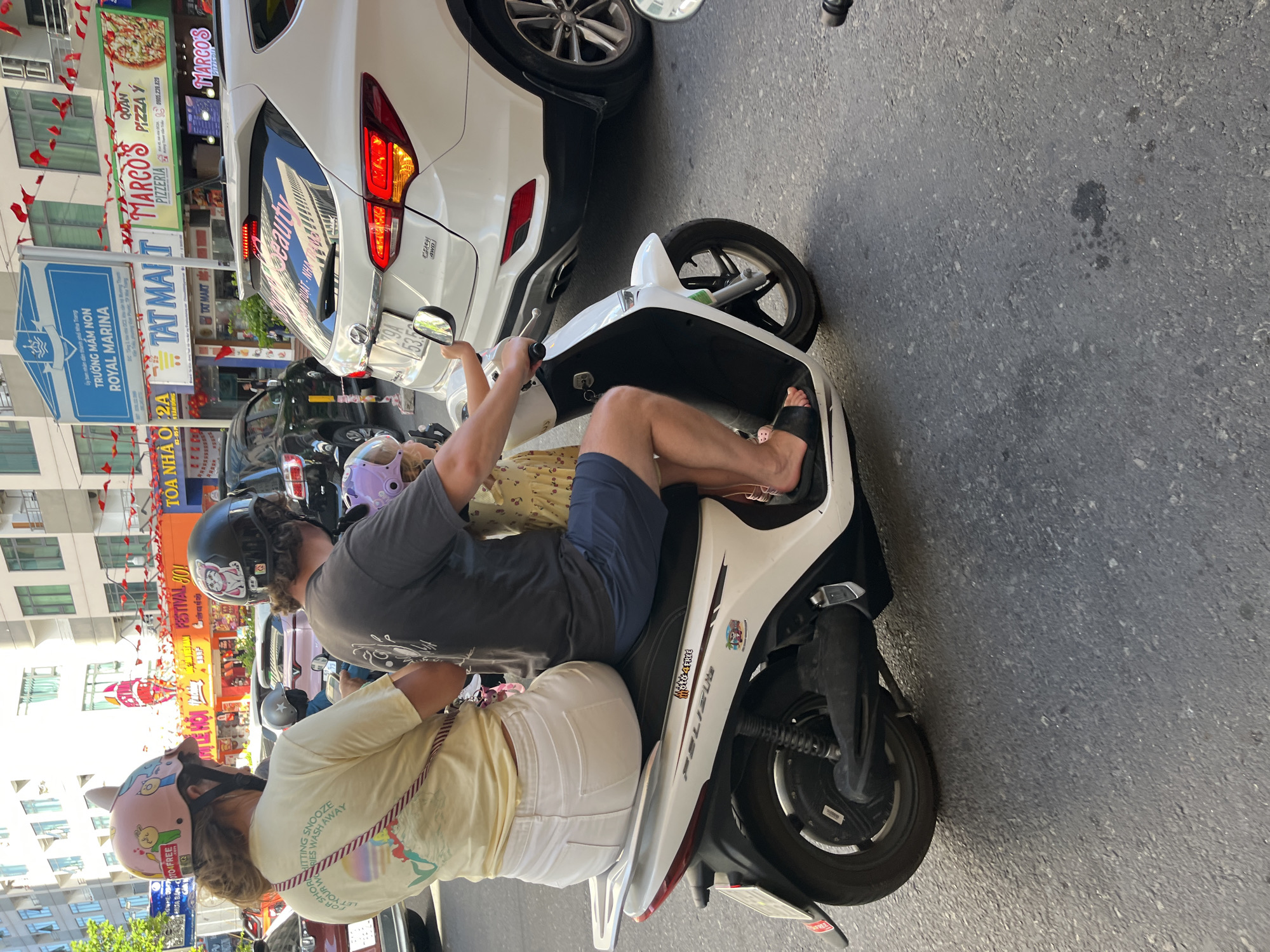 The image shows a family of three riding on a white FELIU electric scooter amidst street traffic in Bac Nha Trang Ward, Vietnam. The driver, wearing a gray shirt, navy shorts, a black helmet, and black sandals, operates the scooter. Behind him sits an adult female wearing a yellow shirt, white pants, and a pink helmet with cartoon characters. A child in a yellow dress and light purple helmet sits between the two adults. The scooter is positioned next to a white car and is surrounded by other vehicles. Behind the scooter and car, a row of shops and restaurants can be seen, including Marco's Pizzeria and a establishment with the text "FESTIVAL JOI." Signs for "TRƯỜNG MẦM NON ROYAL MARINA" and "TAT MAIT" are also visible. Red flags are hung along the street. The car appears to be a Hyundai, with its license plate partially visible.