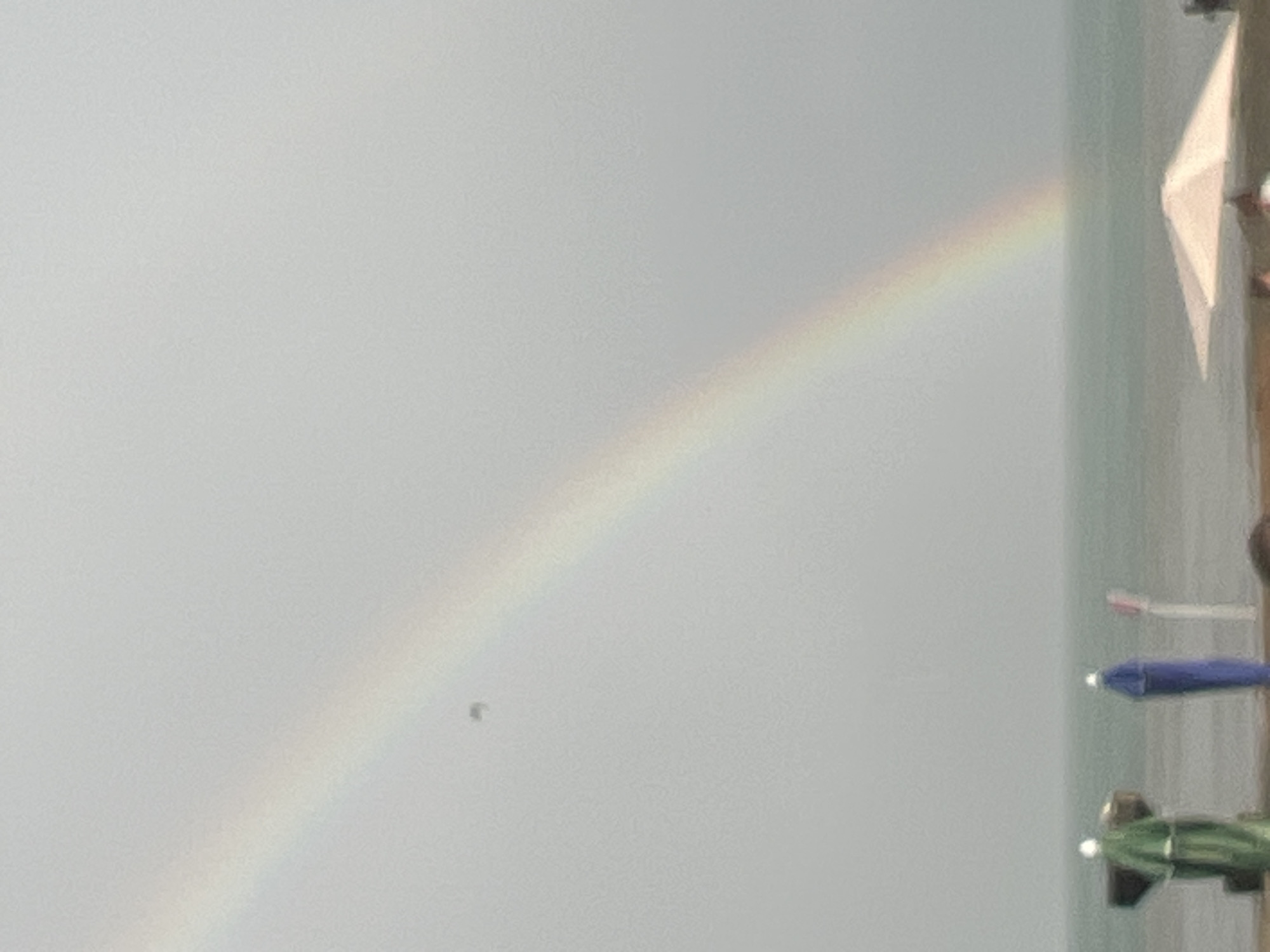 A rainbow arcs across a grey sky over a calm sea. In the foreground, on a beach, are two colorful umbrella-like structures, one blue and one green, and a white umbrella. A single dark object, possibly a bird or drone, is visible in the sky. The scene appears to be a coastal area in Sokhumi, Abkhazia.
