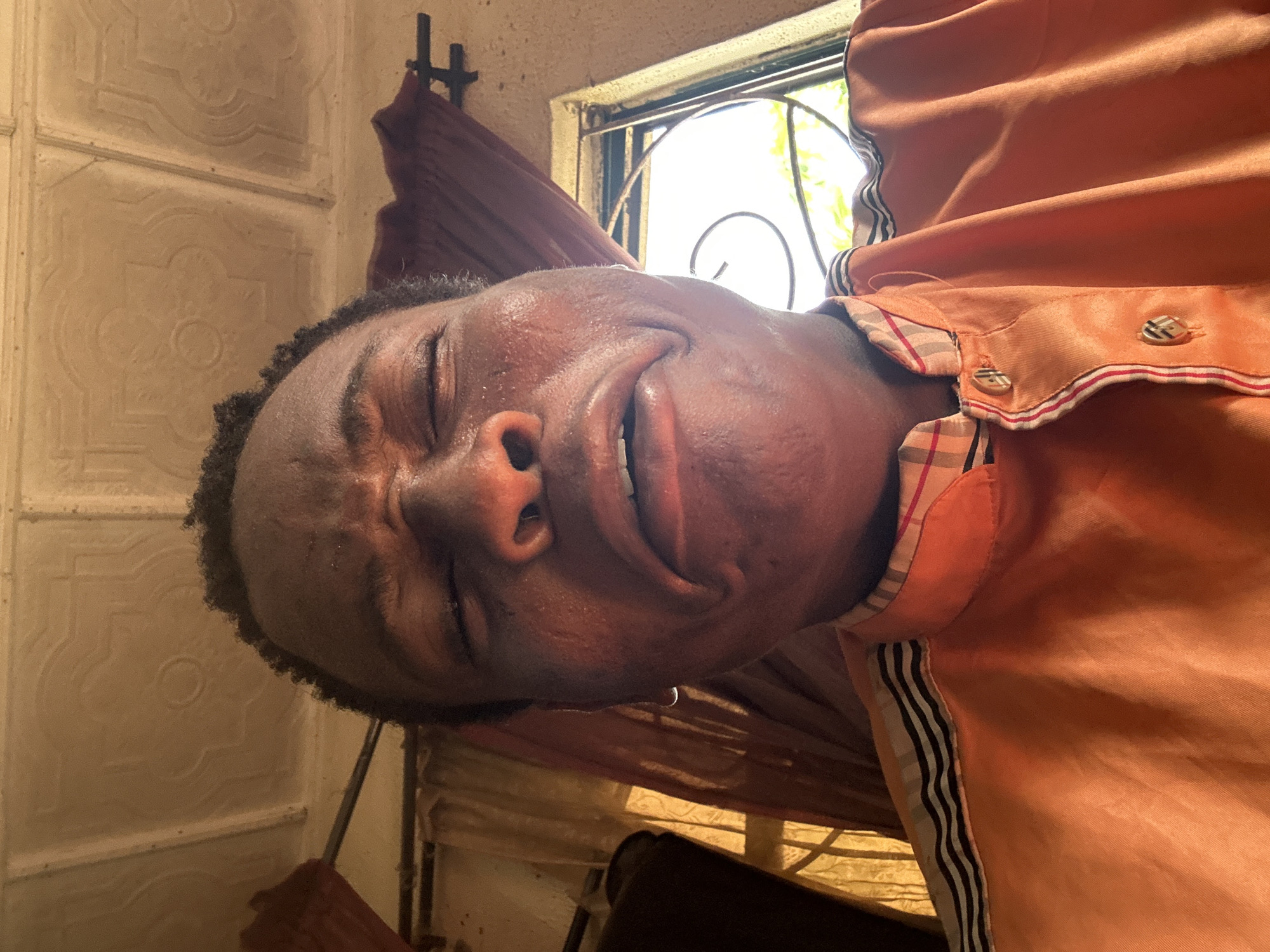 A rotated close-up portrait shows a person, likely male, with dark skin and short, dark, curly hair. The image is oriented with the person's head tilted significantly to the left. Their eyes are closed tightly, and their mouth is open wide, revealing teeth in what appears to be an intense expression, possibly of laughter, crying, or exertion. The person's face glistens, suggesting it is wet or sweaty.
The individual is wearing an orange collared shirt. A plaid pattern is visible on the inside of the collar and on the placket, which features two buttons also adorned with a plaid design. Black and white striped detailing runs along the side of the shirt.
The background includes a light-colored wall with an embossed or patterned texture visible to the left of the person's head. Behind the person and to the right, a bright window allows natural daylight to illuminate the scene, showcasing black metal bars or a decorative grille. Some green foliage is visible through the window. A reddish-brown curtain or piece of fabric is draped below the window. The overall lighting suggests it is daytime. No text is visible in the image.