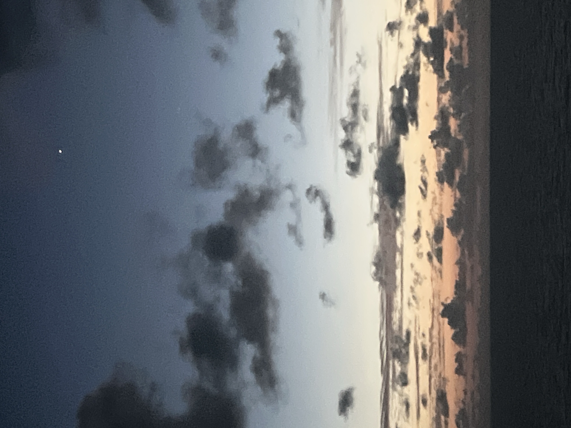 The image depicts a dusky sky over a body of water. The sky is a gradient of colors, starting with a deep blue at the top, transitioning through lighter blues and whites, and ending with soft oranges and pinks near the horizon. Scattered clouds, predominantly dark gray, dot the sky, with some appearing more wispy and others more defined. A small, crescent-shaped moon is visible in the upper portion of the sky. Below the sky, the dark, calm surface of the ocean fills the lower third of the frame. The scene appears to be an outdoor landscape, likely at dusk or dawn, with no discernible urban elements or people.