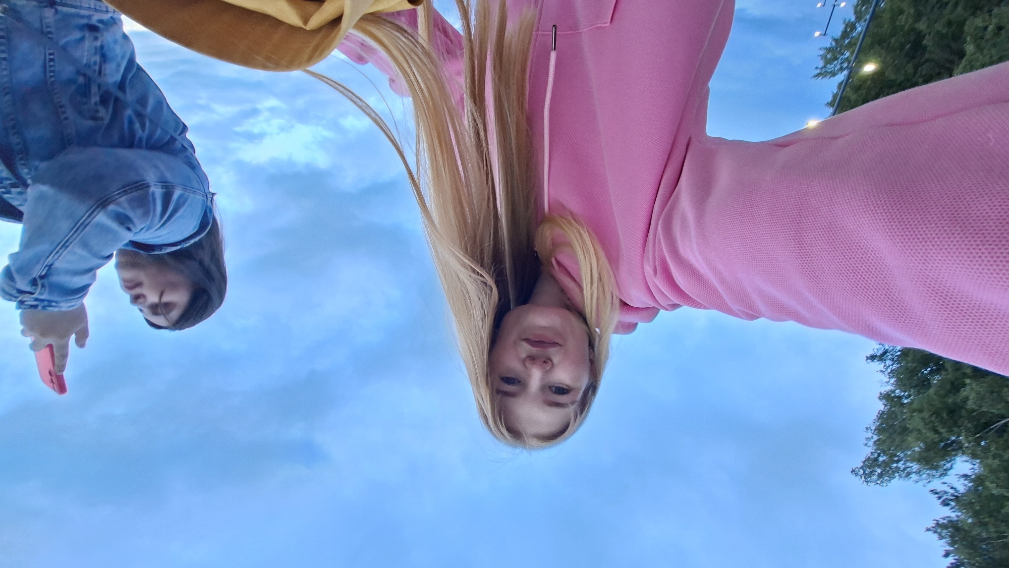 This image, captured in the city of Chaikovsky, Russia, depicts two young women outdoors, viewed from a low angle against a cloudy blue sky.
In the foreground and center-left, a young woman with long, straight blonde hair and fair skin is looking directly at the camera. She is wearing a light pink hooded sweatshirt with a visible textured fabric pattern. Her left arm is extended towards the camera, indicating she is likely holding the device to take a selfie. A white drawstring with metallic tips is visible on her sweatshirt.
To the right, a second young woman with dark hair is partially visible, oriented towards the right side of the frame. She is wearing a light blue denim jacket with visible stitching and texture. Her right arm is raised, holding a bright pink smartphone upwards, suggesting she is taking a photograph or video of something off-frame above. Her gaze is directed upwards and to the right, consistent with her action.
The background consists primarily of a blue sky with scattered white and grey clouds. Dark green tree foliage is visible in the upper left corner and subtly in the bottom left. In the bottom-left quadrant, obscured by the main subject's arm, a light source or streetlamp pole can be faintly seen. A segment of a light brown or tan bag or object is visible in the bottom center, below the blonde woman's right arm.