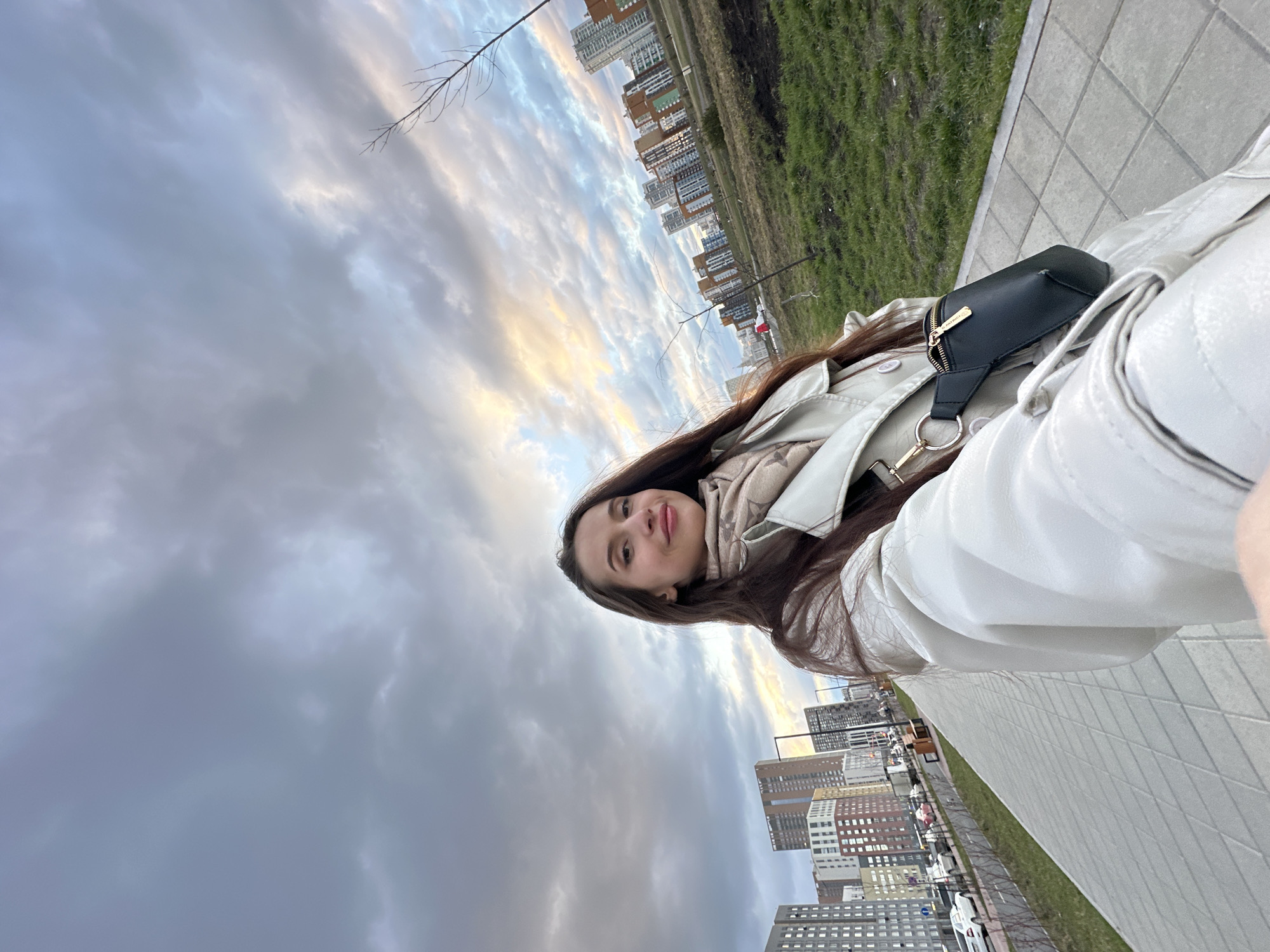 A woman is taking a selfie outdoors. She has long brown hair and is wearing a light-colored jacket and scarf. She also has a small black purse slung over her shoulder. The background shows an urban landscape with tall buildings, a strip of green grass, a sidewalk, and a cloudy sky. The buildings suggest this is in Yekaterinburg, Russia.