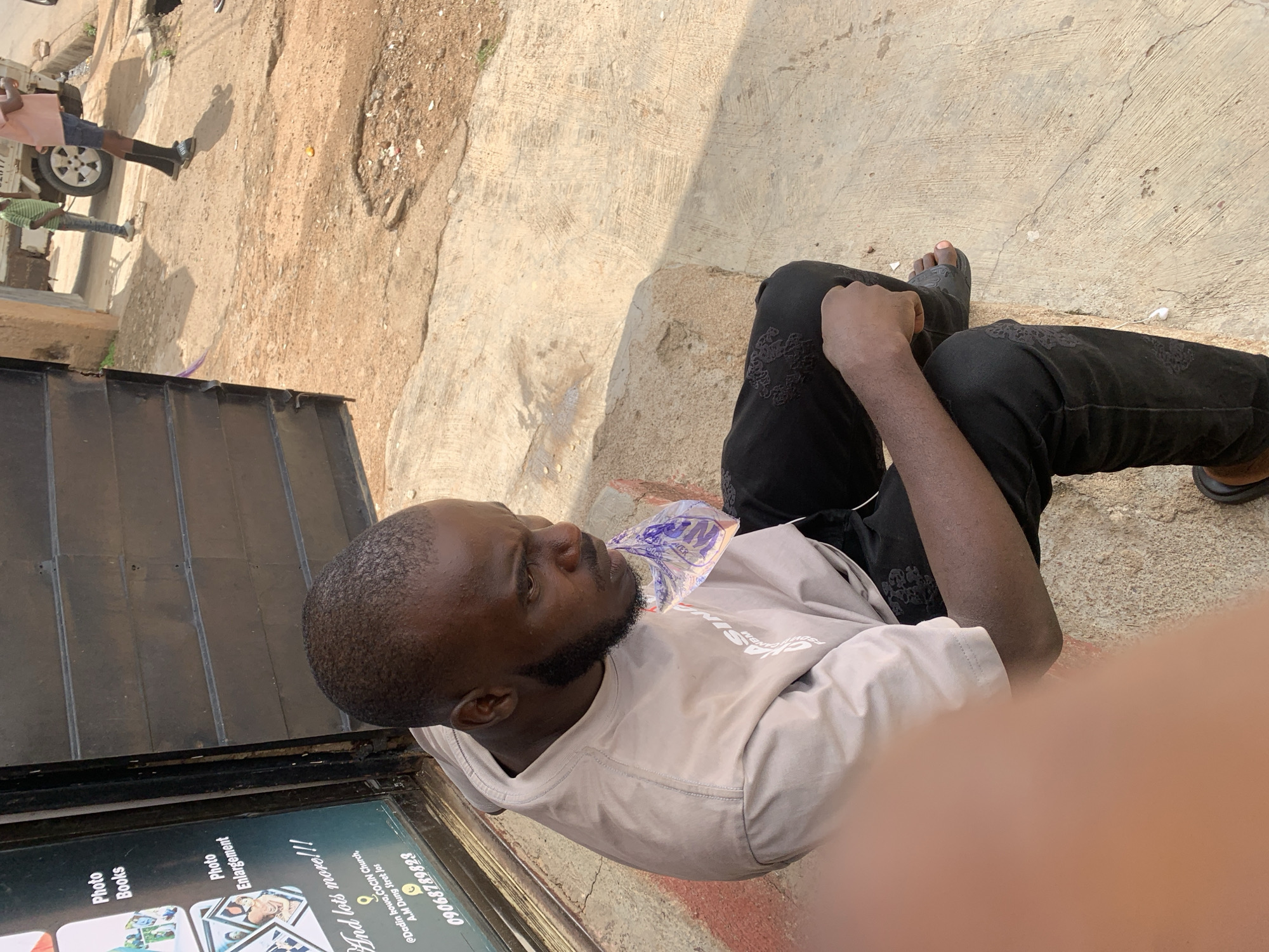 The image, taken in Jos, Nigeria, features a man seated outdoors on a concrete surface, appearing to be taking a break. He is seen with a clear plastic sachet, possibly containing water, near his mouth. The man wears a light grey long-sleeved shirt with some text (partially visible as "C_ASING"), black trousers with an embroidered pattern, and black sandals. He has a short beard and looks away with a neutral, perhaps contemplative, expression.
The setting appears to be an urban or semi-urban street, characterized by a mix of concrete paving and dusty bare earth. In the background, two other individuals are partially visible; one stands near a white pickup truck, and another is further in the distance. The bright sunlight suggests it is daytime, casting clear shadows on the ground, indicative of sunny and dry weather conditions.
On the left side of the frame, there is a dark structure next to a display board. Text visible on the board indicates services offered, such as "Photo Books" and "Photo Enlargement," along with "And lots more!!!". The board also provides a location reference: "@Dadin kowa, COCIN Church, A.M Dung Stret Jos" and a contact number "09068789823". The brand name "ACER" is also partially discernible on the sign.