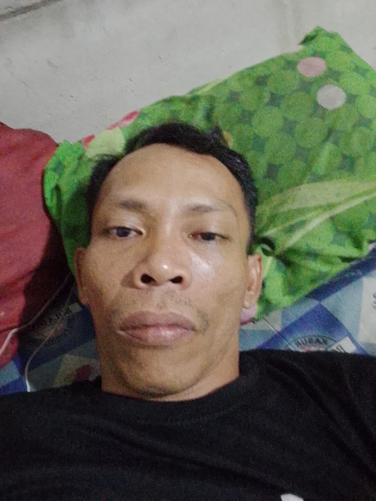 The image is a selfie of a man with short black hair. He has a dark complexion, and his expression is neutral. He is wearing a black shirt. Behind him, there is a green patterned pillow and what appears to be a bed with a blue and white patterned sheet. The background is somewhat blurred, but appears to be an interior setting. It is not possible to ascertain the specific location in Bukit Ketok, Indonesia, or any activity from the image.