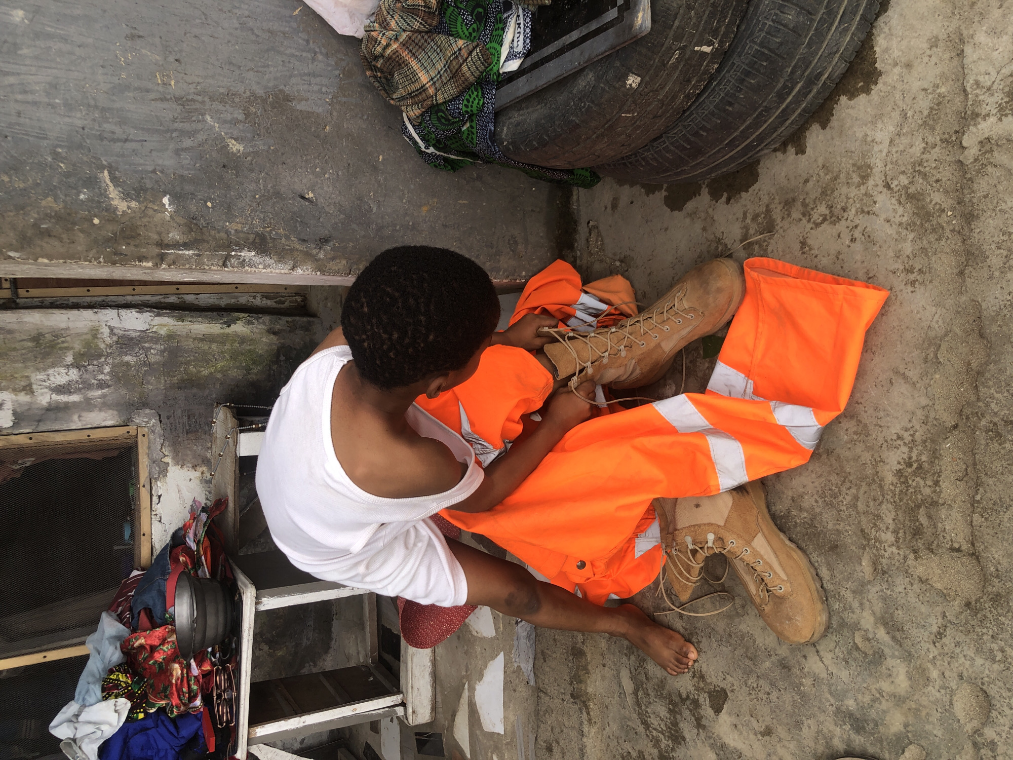 In Ado, Nigeria, a person wearing a white tank top and shorts kneels on a concrete ground. They are in the process of tying the laces of a tan combat boot. In front of them lies a bright orange vest with reflective white stripes, and a second tan combat boot is visible nearby. To the left of the person is a white table with a pile of assorted clothing, and a metal bowl. The background consists of a distressed concrete wall. To the right, stacked tires are piled near some folded patterned fabrics. The ground shows signs of moisture and some scattered debris.