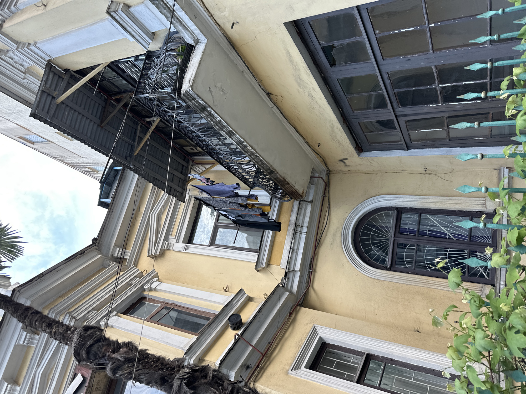 The image, taken from a low angle, showcases an aged building in Batumi, Georgia. The facade is painted in a faded yellow, exhibiting architectural details such as window frames and cornices.
Several windows are visible, including one with a rounded top. From a balcony above, clothes are hung to dry. A rudimentary structure, likely for weather protection, juts out from a higher floor.
The surrounding environment includes a palm tree, visible on the left side, and some green foliage. The image conveys a sense of wear and age, reflecting the architectural style and urban landscape of Batumi.