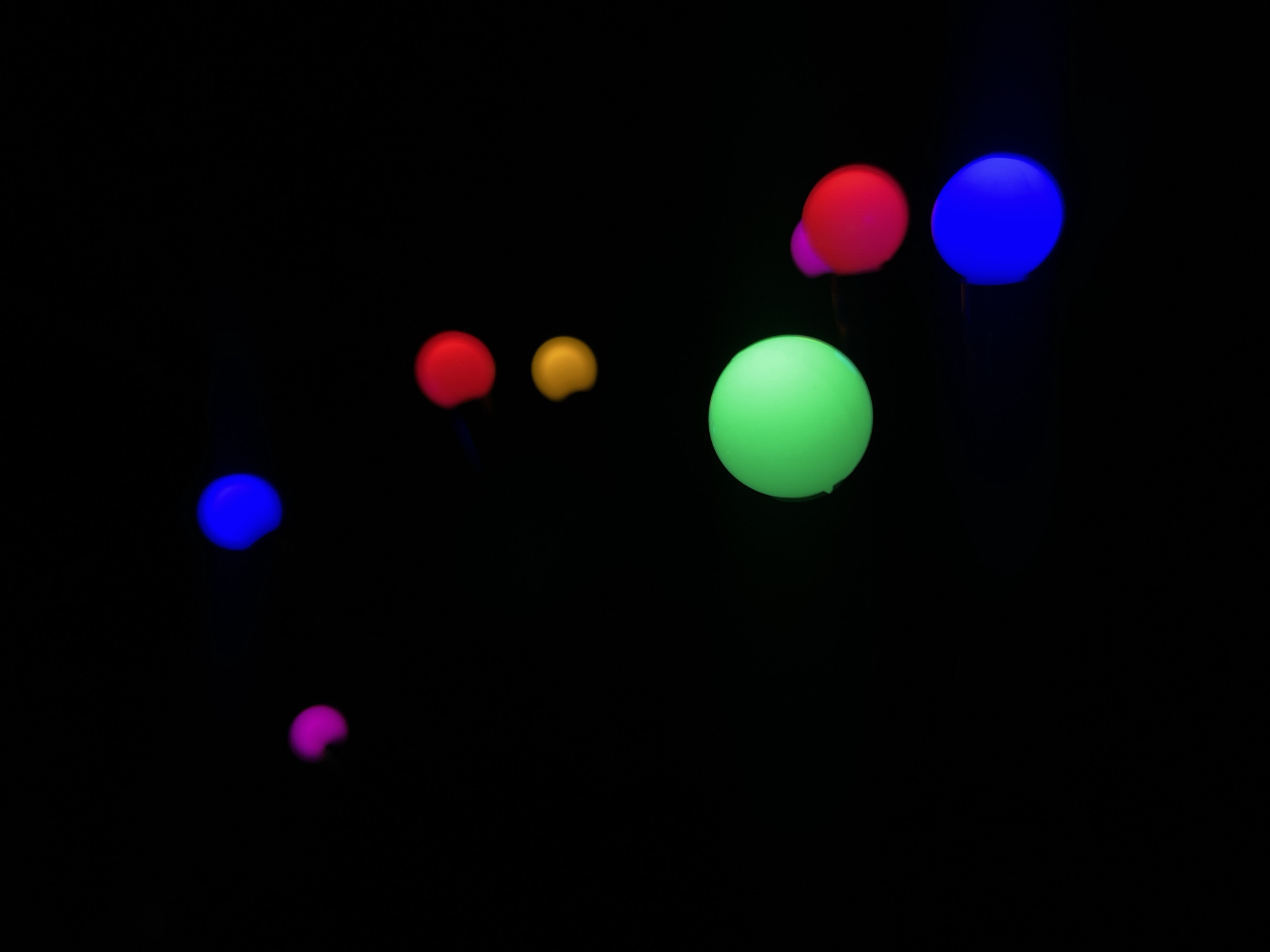 The image shows a black background with several glowing, spherical objects in various colors (green, red, blue, yellow, and pink) scattered across the frame. The objects appear to be light fixtures, likely on poles. The location is unknown, and no people, subjects, or actions are visible. No specific interactions are apparent.