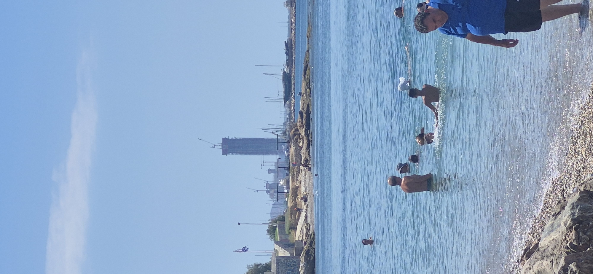 The image depicts a beach scene in Paleo Faliro, Greece, under a clear blue sky with a few wispy clouds. In the foreground, the shallow, clear blue water laps against a rocky and pebbly shoreline. Several people are present in the water, some swimming and others wading.
In the middle ground, a group of people are enjoying the water. One person is standing in the water facing away from the camera, while others are partially submerged. A child is splashing energetically in the water. Further out, a person in a blue shirt and black shorts is walking out of the water towards the shore, looking down.
The background reveals a more distant view of the coastline. A long, thin skyscraper under construction, topped with cranes, dominates the skyline. Other cranes are visible around it, suggesting a large construction project. Several sailboats and yachts are moored in a harbor area. A Greek flag flutters on a flagpole on a small, rocky outcrop to the left. The overall impression is a lively summer day at the beach with ongoing urban development in the distance.