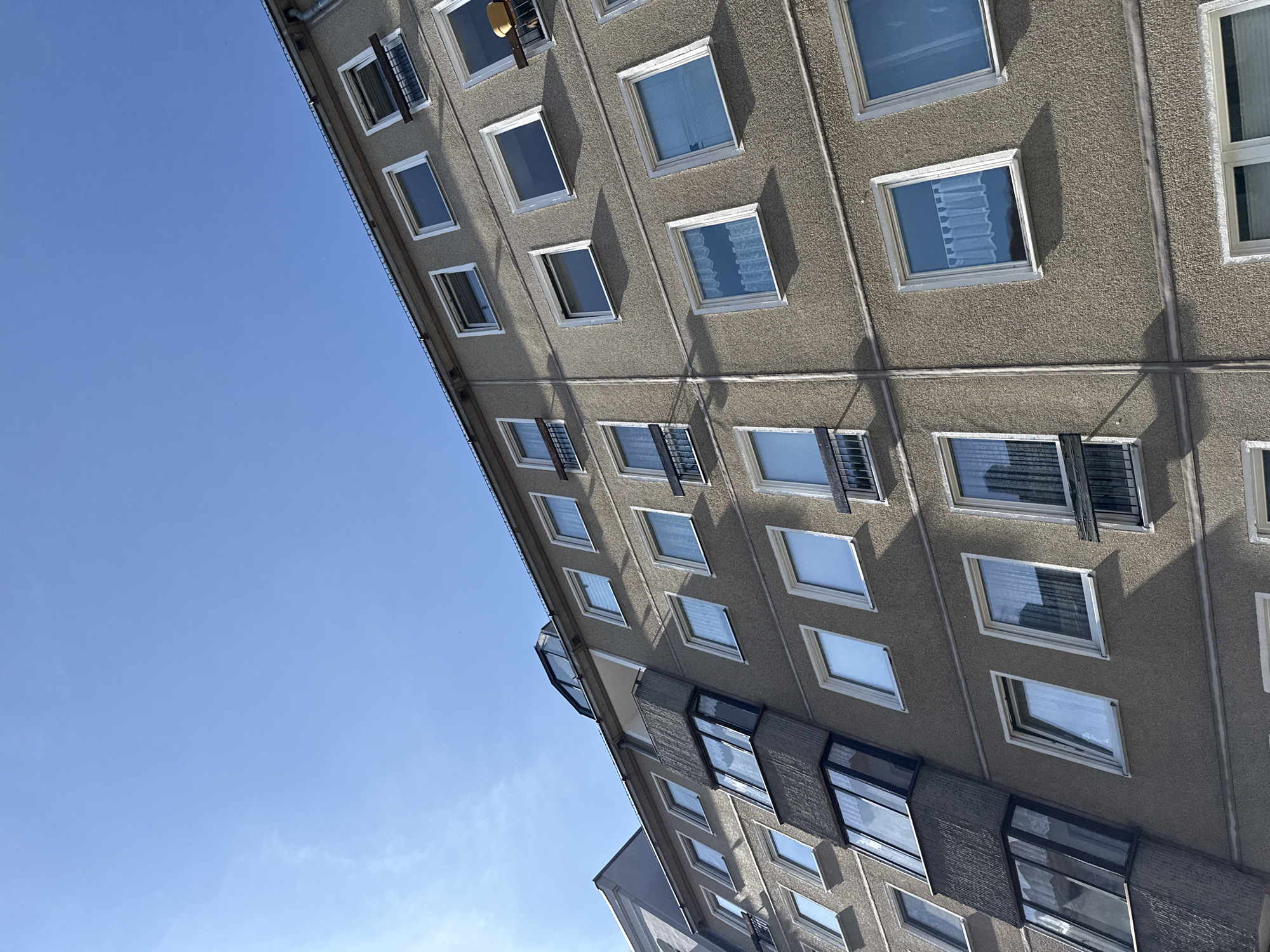 The image depicts a close-up, upward-angled view of a multi-story apartment building facade against a clear blue sky. The building's exterior is a textured, light brown stucco. Multiple windows are visible, arranged in horizontal rows and spaced regularly across the facade. Some windows are open, while others are closed and covered with curtains or blinds, some of which are sheer. Several windows have small, dark balconies or window boxes with railings. A prominent, angled balcony structure is visible on the left side of the building. The building appears to be in an urban setting, possibly in the city of Görlitz, Germany, as suggested by the architectural style often found in post-war German housing. The lighting indicates it is daytime, with sunlight casting shadows on the building. There are no people or discernible objects other than the building itself and its windows.