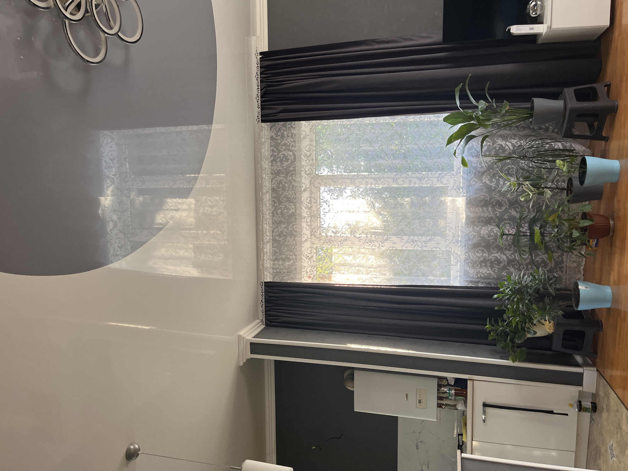 The image depicts an indoor scene, likely a living room or dining area. A large window with white, patterned sheer curtains and dark grey drapes dominates the center of the frame. Sunlight streams through the window, illuminating the room and creating a bright, airy atmosphere. Several potted plants are arranged in front of the window, adding a touch of greenery.
In the foreground, to the left, a white cabinet with a metallic handle is visible, suggesting a kitchen or storage area. Above the cabinet, a white appliance, possibly a water heater, is mounted on a dark grey wall. To the right of the window, a black stool holds a potted plant. Further to the right, a portion of a white entertainment unit with a television is visible.
The ceiling is a striking feature, with a glossy, multi-level design incorporating white and dark grey sections. A modern chandelier hangs from the ceiling on the right side of the frame. The walls are painted in dark grey tones, creating a contrast with the lighter ceiling and window treatments. The floor is made of polished wooden planks.
There are no people visible in the image. The scene appears to be a well-maintained residential interior. Based on the provided information, this scene is located in Saratov, Russia.