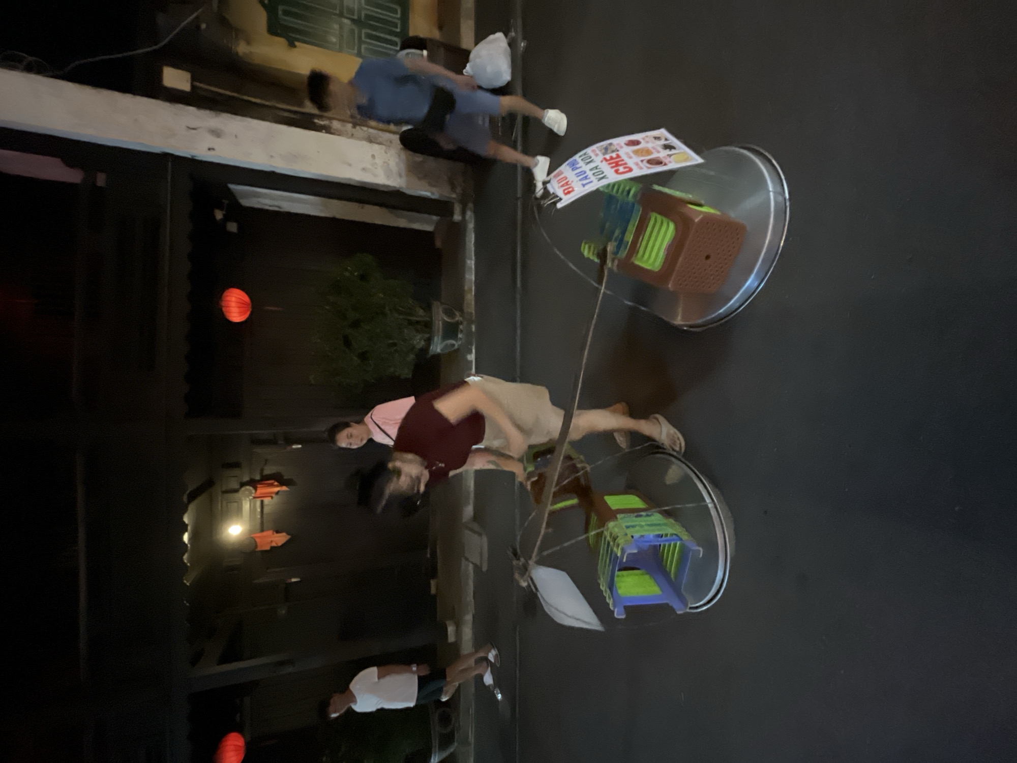 At night on a street in Hoi An, Vietnam, a woman wearing a maroon shirt and beige skirt bends over a portable food cart. The cart has two colorful plastic containers stacked on a metal tray. She is holding a pole attached to the cart, which has a white umbrella on top. To the right of this cart, another identical cart is visible, also with a sign displaying food items. A man in a blue shirt and shorts walks away from the carts, while a woman in a pink shirt stands behind the first cart. Further in the background, a man in a white t-shirt and shorts walks away. Red lanterns hang from a building in the background.