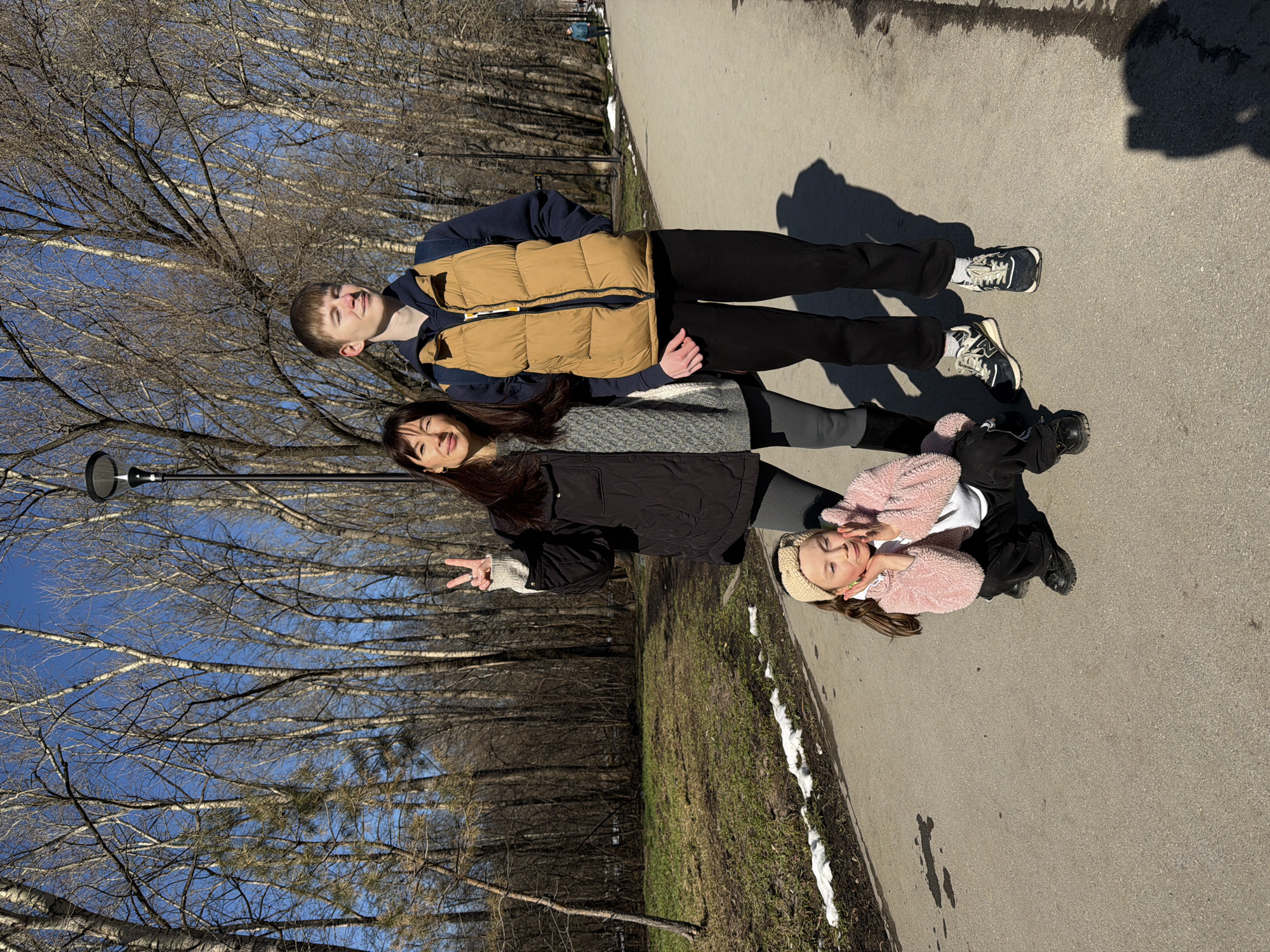 The image shows three individuals lying on a paved pathway in a park-like setting, possibly in the city of Perm, Russia, given the presence of bare trees characteristic of the region's climate. A young man at the top wears a tan puffy vest over a dark shirt, dark pants, and sneakers. A woman is in the middle, dressed in a dark jacket over a grey patterned top and dark leggings. She is making a peace sign. A young girl at the bottom wears a pink fuzzy jacket, a white top, dark pants, and a cream-colored knit hat. The pathway is bordered by grass and some remnants of snow. A line of bare trees and a blue sky with some light cloud cover is visible in the background. A lamppost is seen on the left side.