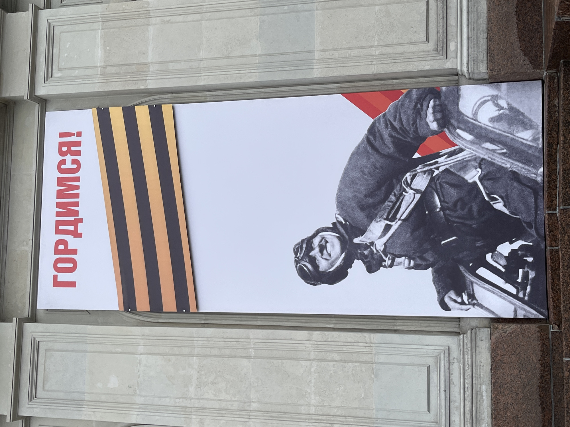 A rectangular banner is affixed to a building facade in Saint Petersburg, Russia. The banner features the text "ГОРДИМСЯ!" (We are proud!) in red, followed by a black and orange ribbon pattern. A black-and-white image of a person wearing goggles and gear is shown riding a motorcycle. The building facade is constructed of gray stone with recessed panels. To the right of the banner, the building is constructed of a darker, mottled stone.