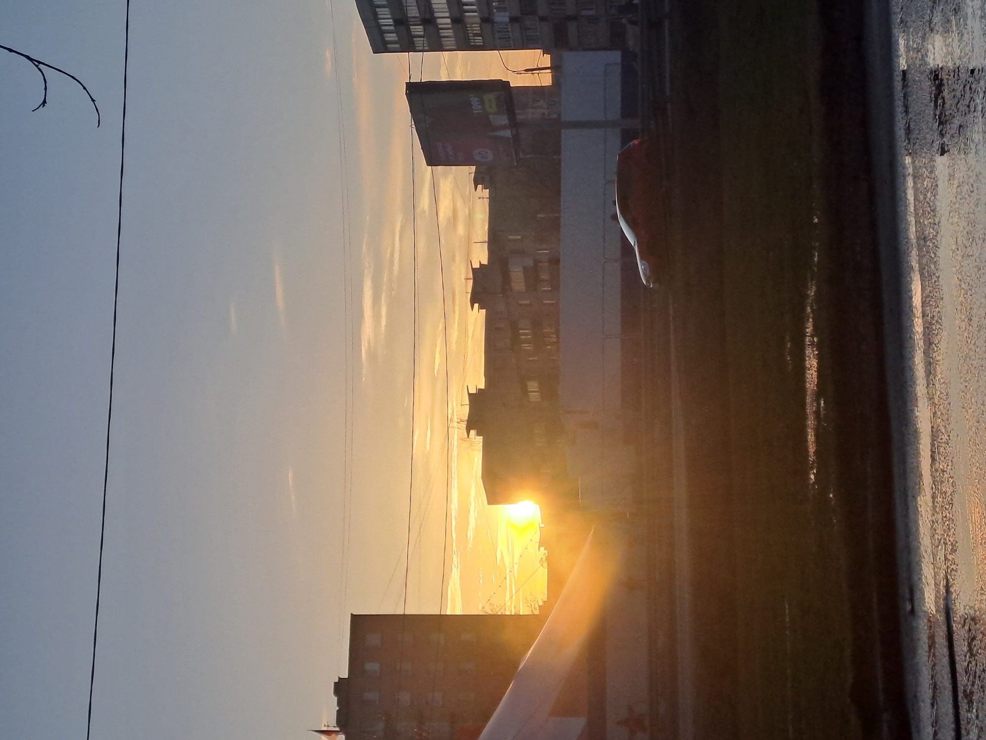 The image captures a sunset in what appears to be an urban area. Buildings are silhouetted against the bright setting sun. The presence of a billboard suggests a commercial or residential district. Reflections visible in what seems to be a wet surface in the foreground indicate a possible rain or condensation. Given the prompt specifies Elektrostal, Russia as the location, the urban landscape likely corresponds to the architecture and infrastructure typical of this city. Details such as the specific types of buildings, signage on the billboard, or the precise make and model of the reflective surface's object are not clear, limiting the forensic analysis.