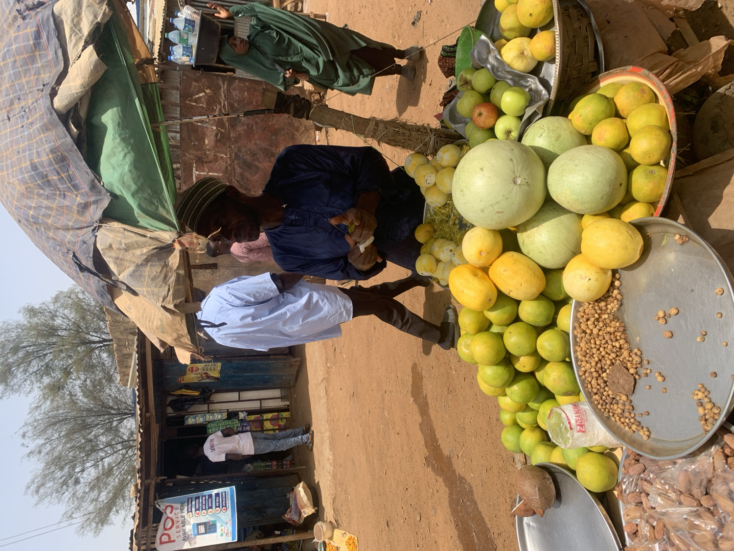 This image captures a vibrant, sunlit scene at a roadside market stall in Rafin Par, Nigeria. The setting is an unpaved, dusty area with simple, makeshift structures, including a tent-like canopy fashioned from tarps and a small, basic shop building with a corrugated metal roof.
Several individuals are present, engaging in daily activities. A central figure, a man wearing a dark blue garment and a green cap, is seen interacting with the market goods. Another man in a light blue shirt stands nearby, while a person in a white t-shirt is visible further back near the shop entrance. In the distance, another person in a green robe is also present.
The primary focus of the scene is a colorful display of fresh produce arranged on metal trays and in woven baskets. These include large light green melons, numerous yellow and green citrus fruits (likely lemons, limes, or grapefruits), and a few red and green apples. On separate metal trays, small, light brown pebble-like items, possibly dried seeds or nuts (like tigernuts), and larger dried fruits or nuts are laid out for sale. A single coconut rests on another tray. Stacks of bottled water are visible behind the central vendor.
The bright sunlight and strong shadows indicate it is daytime, likely mid-morning or afternoon. The overall impression is one of a bustling local marketplace. Visible text on a blue sign near the shop reads "POS SERVICES AVAILABLE HERE" along with icons suggesting mobile banking or payment services.