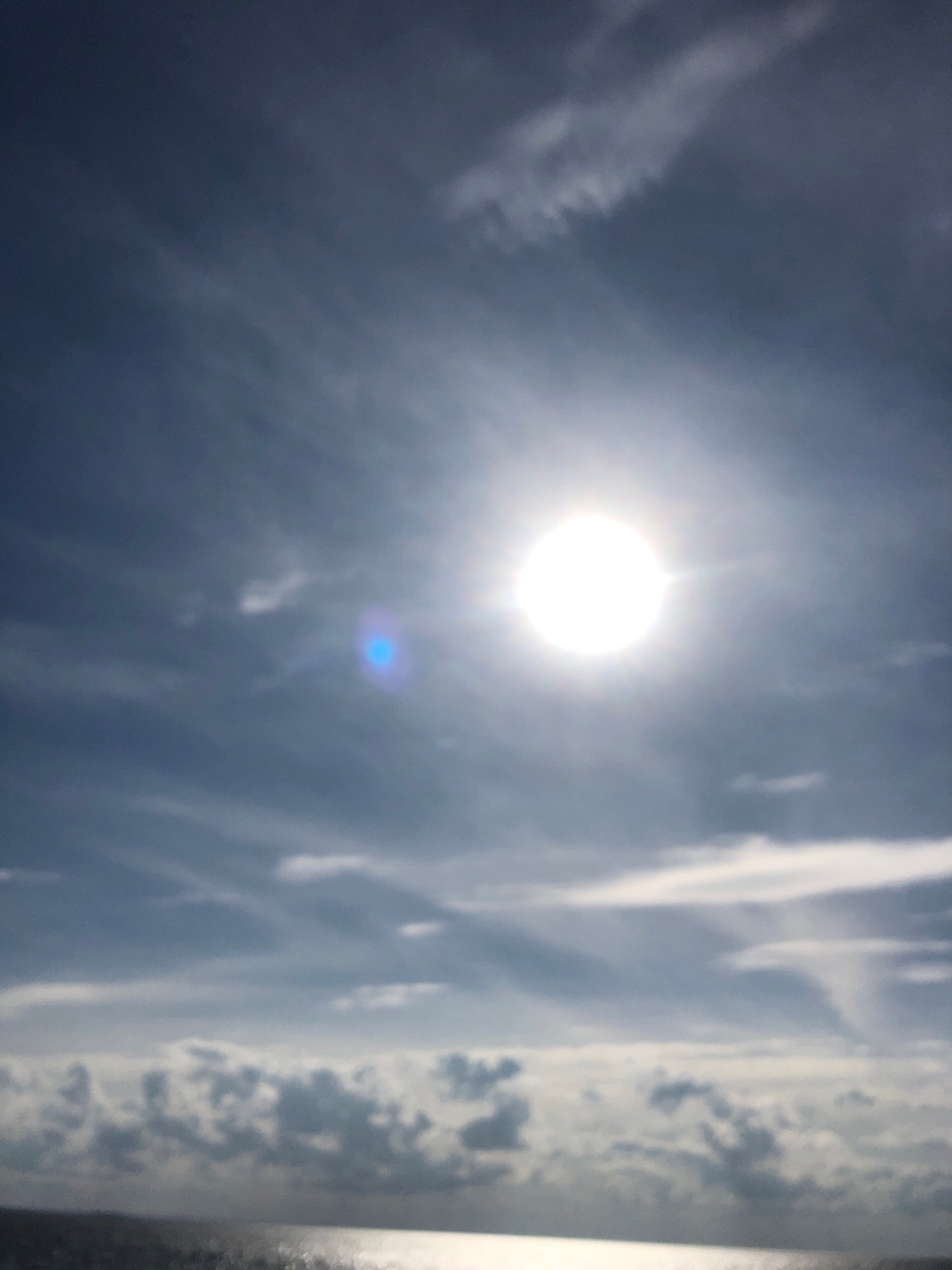 The image depicts a bright, sunlit sky over a body of water. The sun is prominently visible, appearing as a large, luminous orb in the upper center of the frame. Wispy clouds streak across the deep blue sky, creating a textured atmosphere. A distinct lens flare, appearing as a bright blue and white halo, is visible to the left of the sun. In the lower portion of the image, the surface of the water is visible, reflecting the sunlight and appearing somewhat turbulent or rippled. The overall scene suggests an outdoor environment, possibly a seascape or waterscape, with a clear and bright day. There are no people or specific man-made objects clearly identifiable. The image appears to be taken from a low vantage point, looking towards the horizon.