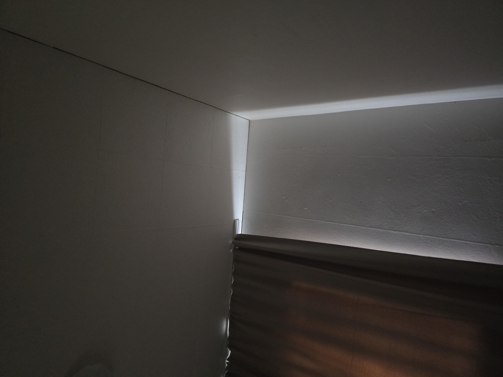 The image shows a corner of a room where the ceiling meets two walls. A curtain hangs on the left side of the frame, obscuring a portion of the wall. The ceiling is textured, with visible seams. A bright light emanates from the junction of the ceiling and the wall, creating a dramatic highlight. The walls are also textured. The scene is dimly lit, with shadows cast by the curtain and the angle of the light. There are no people or animals visible in the image. The location of Chaplynka, Ukraine is not discernible from the image itself.
