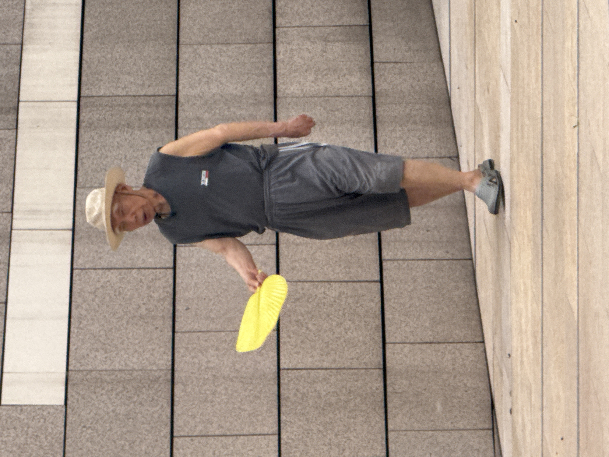 An elderly East Asian man wearing a wide-brimmed straw hat, a sleeveless dark gray t-shirt, and gray shorts walks on a light-colored tiled pathway. He holds a bright yellow fan in his right hand, extended slightly. His left arm is bent at the elbow, and his hand is at his side. The man is wearing dark gray slip-on shoes. Behind him is a textured wall made of rectangular stone tiles, alternating between lighter and darker shades of gray. The scene appears to be outdoors in an urban environment. The man's posture suggests he is walking leisurely. The lighting indicates it is daytime.