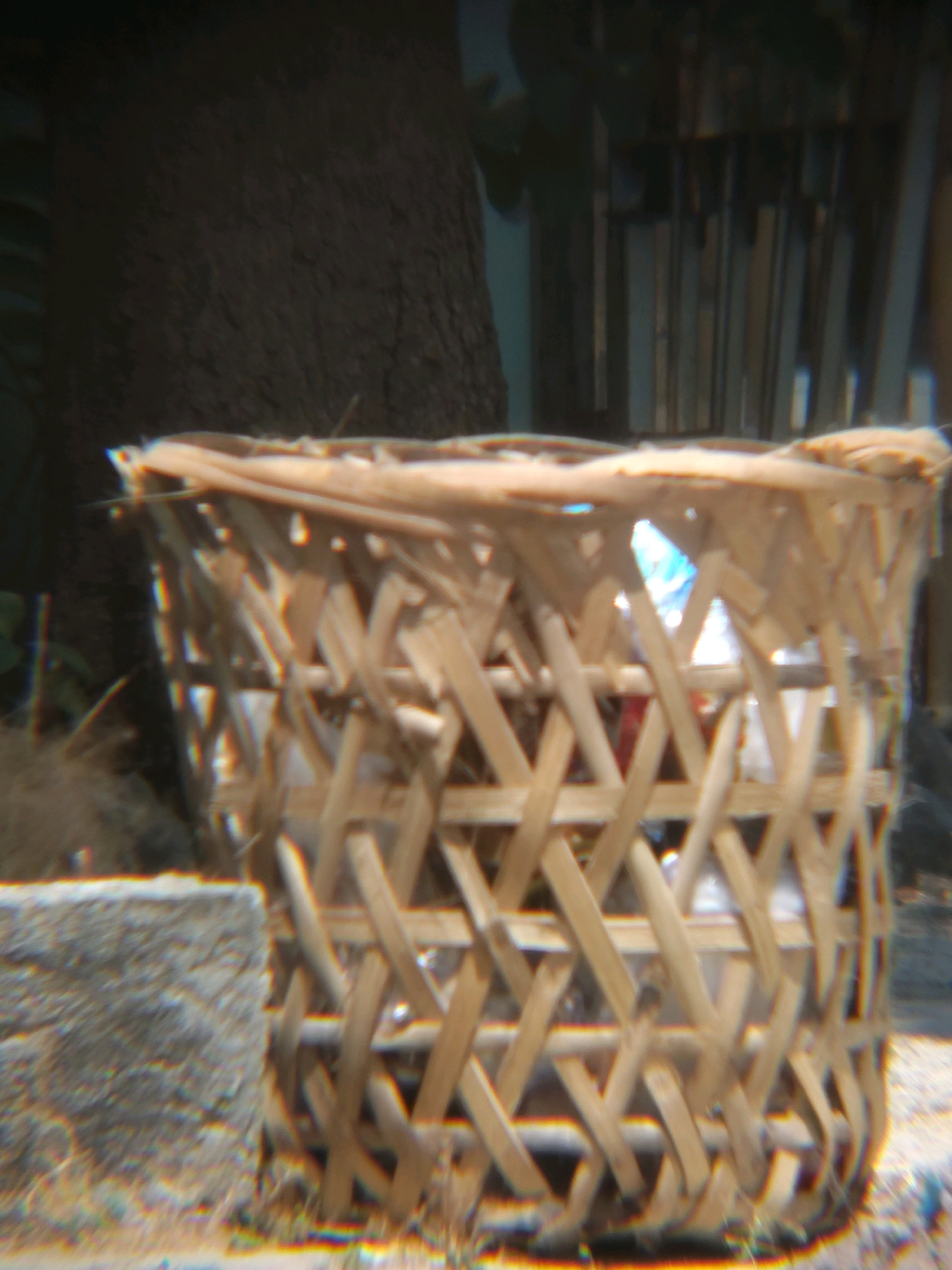 In the city of Bulak Lor, Indonesia, a close-up, slightly blurry image captures a woven bamboo basket overflowing with translucent plastic bags. The basket, made of light-colored bamboo strips interwoven in a crisscross pattern, sits on what appears to be the ground, possibly sand or gravel. To the left of the basket, a rough-textured, rectangular stone or concrete block is visible. The background is out of focus, but suggests a dark, possibly wooden structure or a tree trunk, with vertical elements that could be fences or stacked lumber. Light filters through the basket, illuminating the plastic bags within.