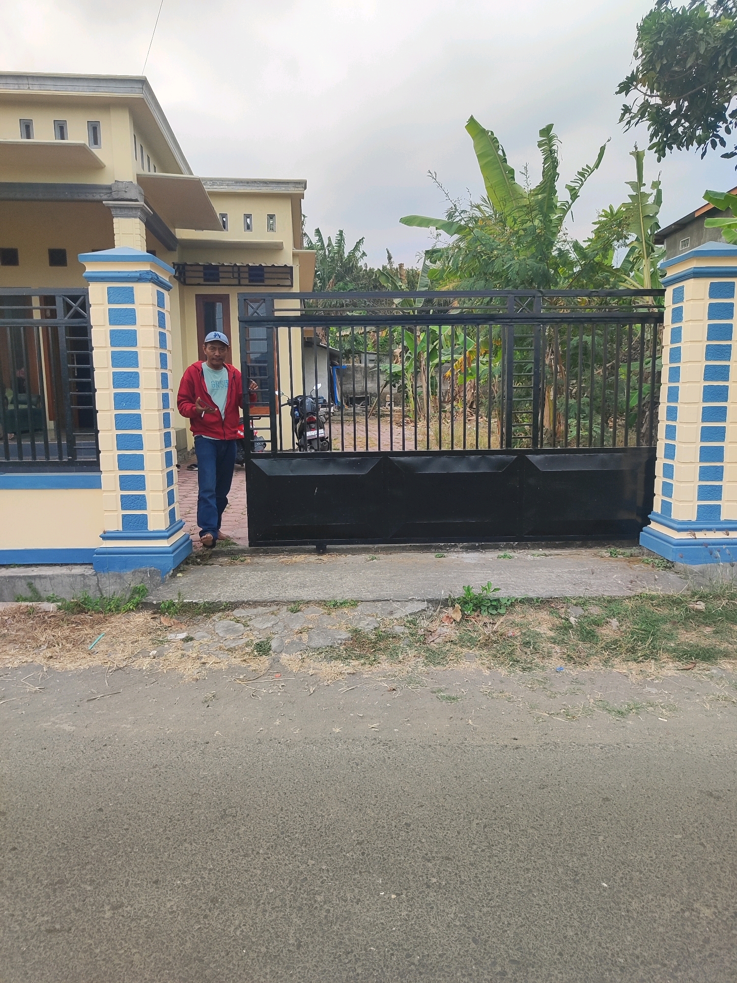 A man is standing in front of a black metal gate and a light yellow house with blue and yellow striped pillars. He is wearing a red hooded jacket, a light blue t-shirt, blue jeans, and a blue baseball cap. He appears to be holding something in his right hand. Behind the gate, there are various trees and plants, including what appear to be banana trees. A motorcycle is visible parked behind the gate to the right of the man. The scene is outdoors with a paved road in the foreground and a cloudy sky above. The location is identified as Rejowinangun, Indonesia.