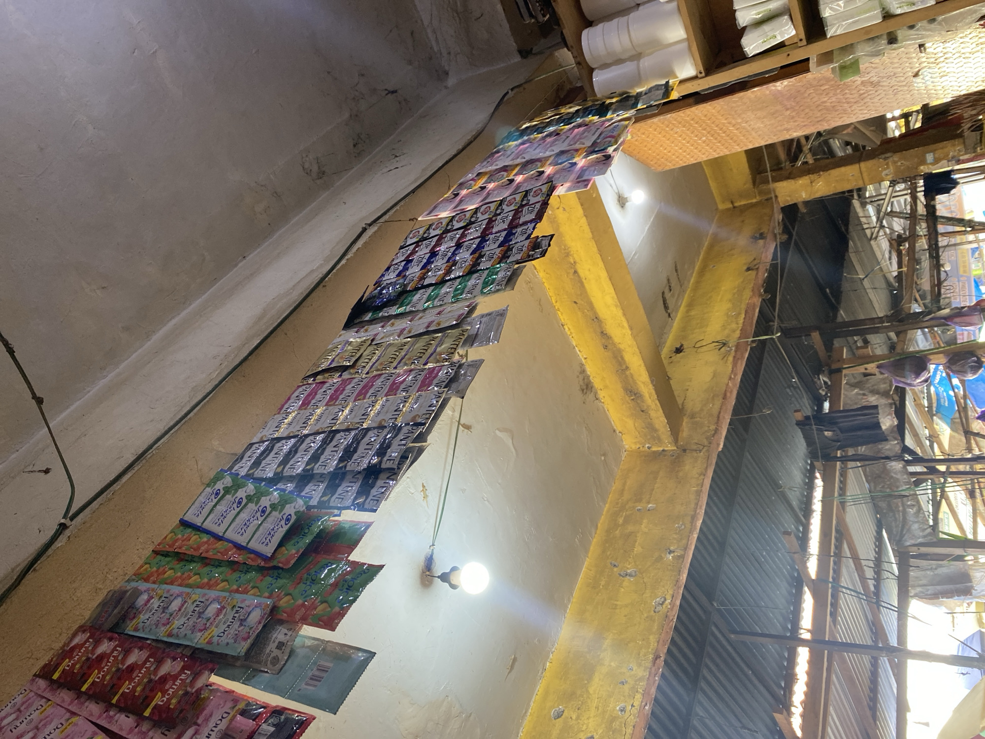 In the city of Ta', Indonesia, within a dimly lit market stall, an assortment of small, colorful sachets of personal care products, likely shampoo and conditioner, are neatly arranged and displayed. These sachets, featuring various brands and colors, are hung from wires suspended from the ceiling, creating a visually dense and organized presentation. A single bare light bulb illuminates the space, casting a warm glow on the products and the surrounding utilitarian structure of the market, characterized by painted yellow beams and a corrugated metal roof. The scene depicts a common retail environment where small, individually packaged items are showcased for sale, suggesting a bustling local marketplace.