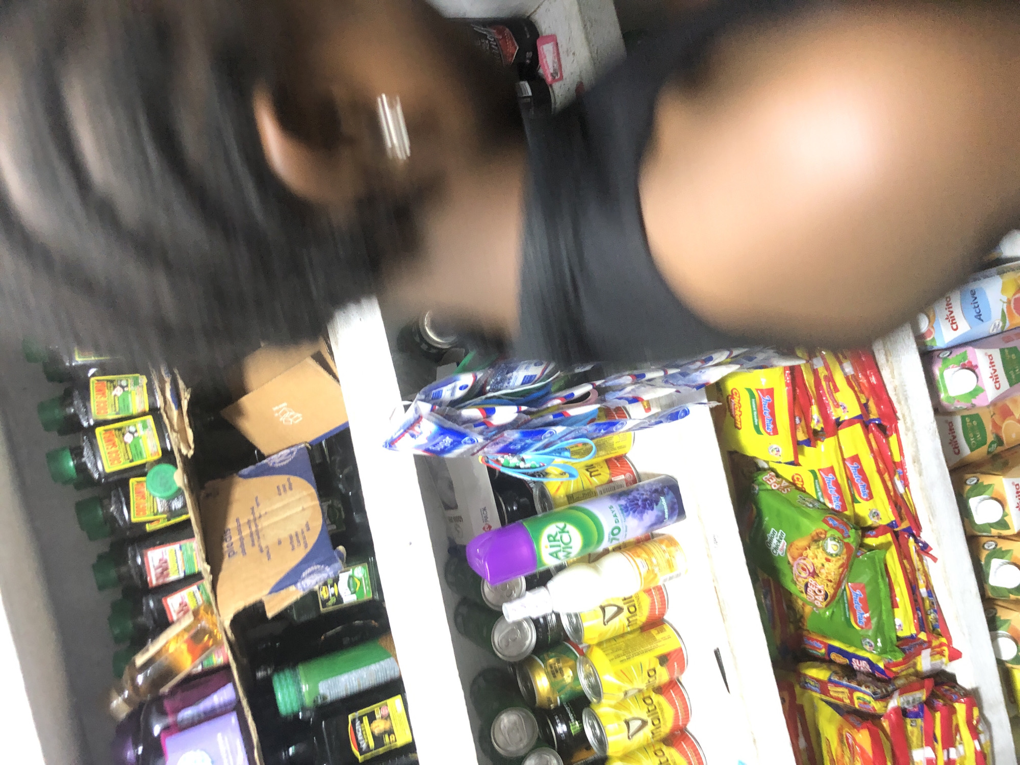 The image shows a shelf in a store, likely a small convenience store or kiosk, in Ado, Nigeria. The shelf is stocked with various items, including beverages in cans and bottles, as well as packaged food items. Among the beverages are cans of Guinness Malta, and some yellow-labeled cans that are difficult to identify. There is also a bottle of "Air Wick" lavender air freshener. Packaged food items, appearing to be instant noodles, are stacked on a lower shelf. The background is blurred, but it is possible to see a person's shoulder and hair, suggesting a customer or employee is in the vicinity. The lighting is dim and appears to be artificial, typical of an indoor environment.