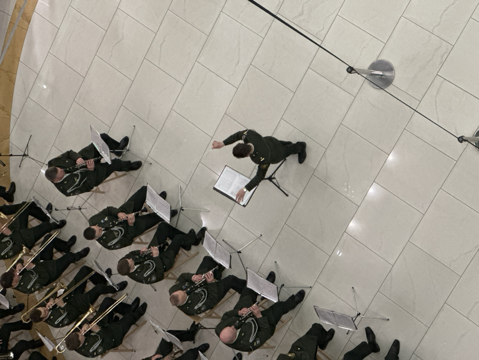 An overhead view captures a male military-style brass and woodwind band performing indoors, likely within a modern public space such as a shopping mall atrium in Kyiv, Ukraine. Approximately fifteen musicians are seated on light-colored wooden folding chairs, playing trombones and clarinets from sheet music displayed on portable stands. All members wear dark green uniforms adorned with light-colored shoulder cords and insignia. A conductor, similarly uniformed, stands facing the ensemble, actively directing with both hands raised. The performance takes place on a floor composed of large, light-grey rectangular tiles. A silver stanchion with a black rope barrier is partially visible on the right side of the frame.