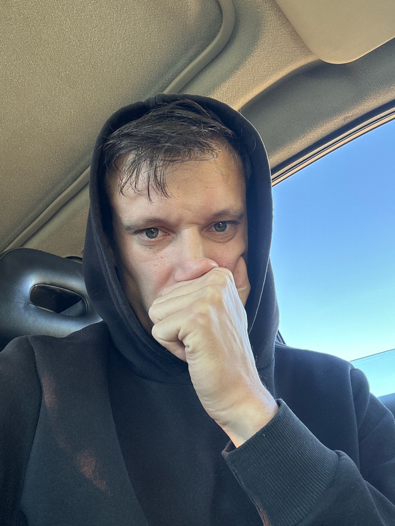 A man is sitting in a car, wearing a black hoodie. He is looking at the camera with his hand covering his mouth. The sky is visible through the car window. The image was taken in Makiv, Ukraine.