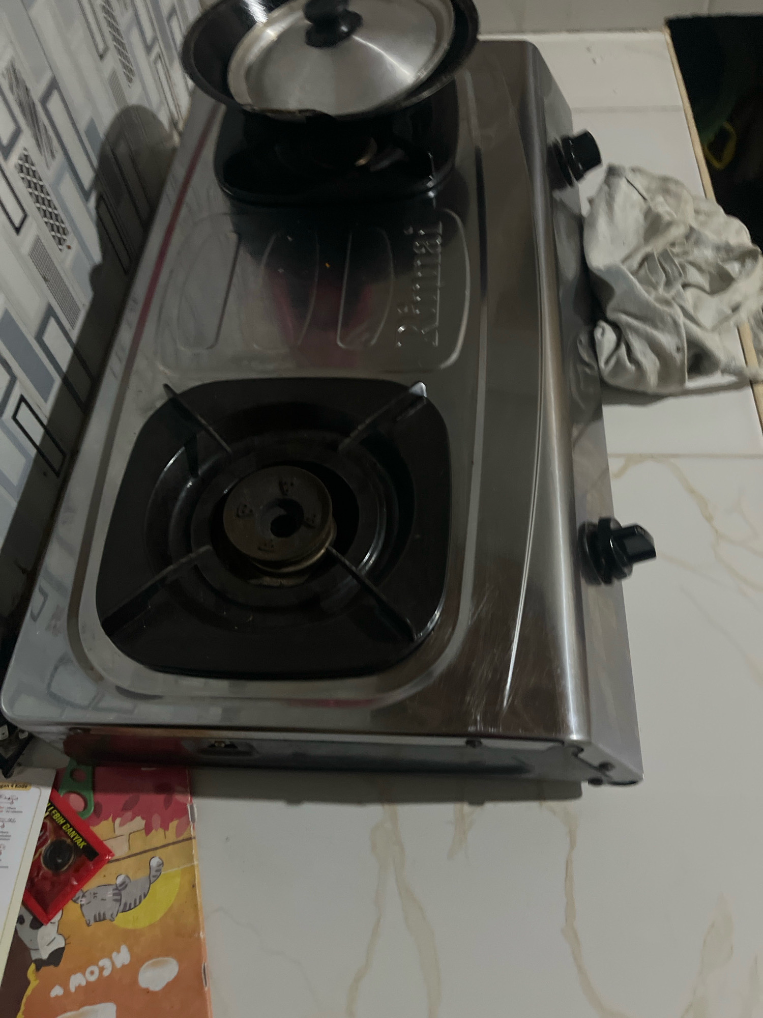 A stainless steel two-burner gas stove sits on a kitchen counter with a marble-patterned laminate finish. A black pot with a silver lid rests on the left burner, which is turned off. To the right of the stove, a crumpled grey cloth is draped over the counter. In the background, a tiled wall features a geometric pattern of grey and white rectangles. In the foreground, a colorful children's book with cartoon cats and the word "meow" is partially visible, along with a red packet and what appears to be a sheet of paper with text and numbers. The scene is illuminated by overhead lighting, casting shadows on the wall. The overall impression is one of a functional, everyday kitchen setting.
