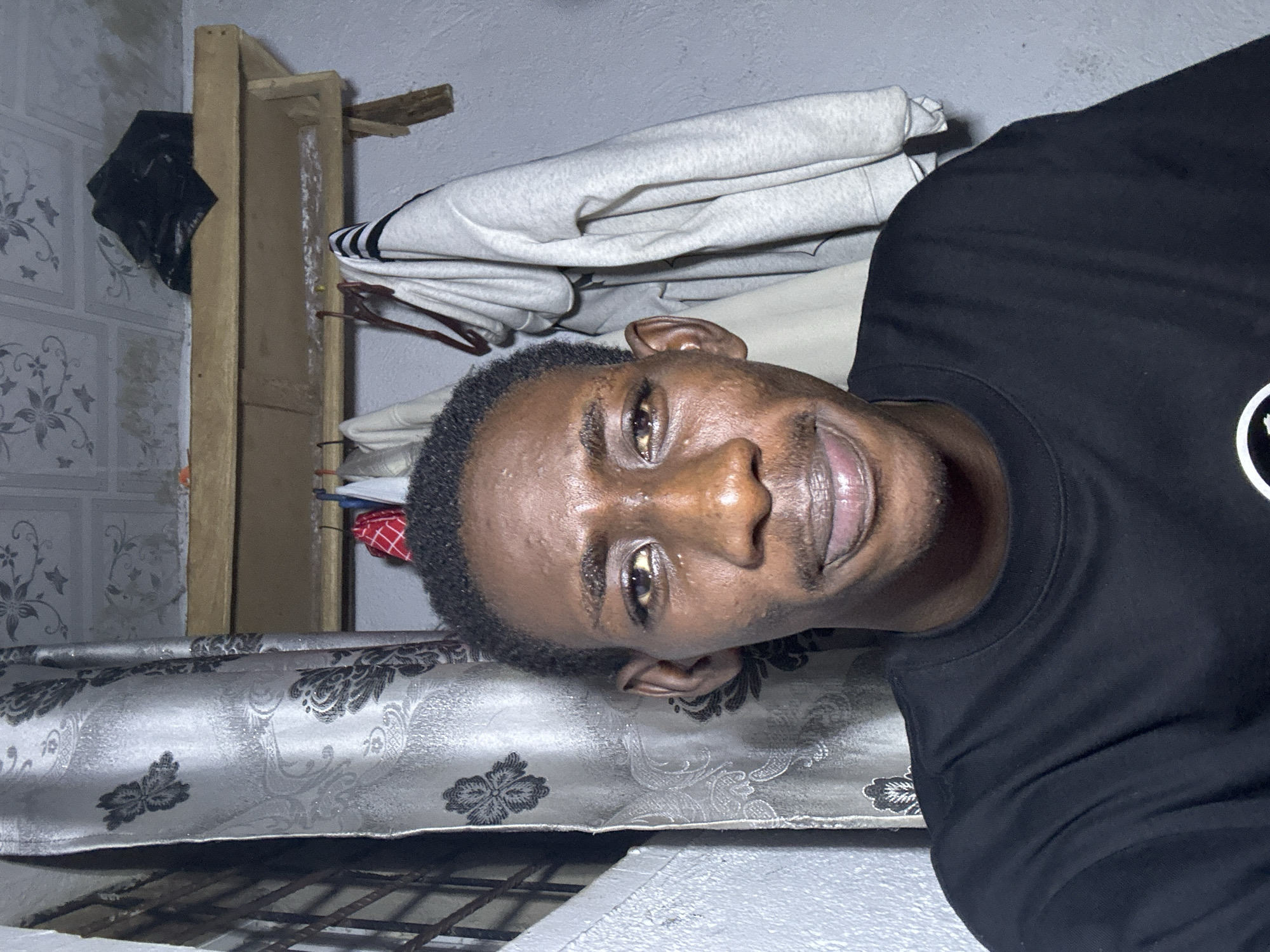 A young Black male is captured in a close-up, waist-up selfie. He has short, dark hair and is wearing a black t-shirt. His face is turned towards the camera, and he has a subtle smile. The background suggests an indoor setting with a patterned curtain on the left, a wooden shelf with clothes hanging on it, and a ceiling with a textured pattern. The lighting is somewhat dim, casting slight shadows. This scene is located in Durumi, Nigeria.