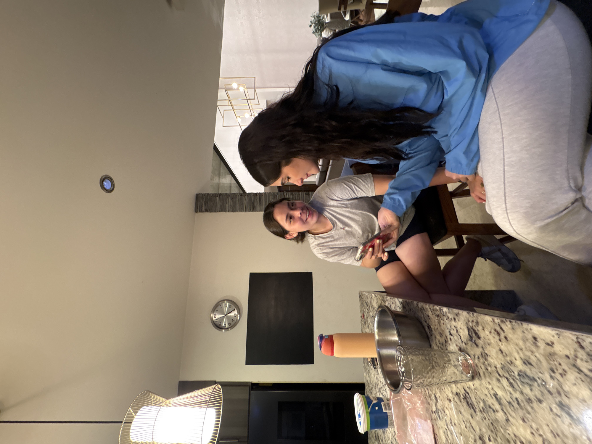 Two women are captured in a casual indoor setting, likely a modern kitchen or dining area in San Bernardino Tlaxcalancingo, Mexico. The image is rotated 90 degrees clockwise.
In the foreground, a woman with dark hair pulled back, wearing a grey t-shirt and dark shorts, is seated on a chair. She is smiling broadly and looking up towards the second woman. Her hands are holding a smartphone, seemingly engaged with its screen.
Leaning over her, from the right side of the frame, is another woman with long, dark hair. She is dressed in a light blue jacket or sweatshirt and light grey sweatpants. Her gaze is directed downwards, suggesting she is interacting with the first woman or looking at the phone she is holding. Their proximity indicates a close and friendly exchange.
The background reveals a well-lit space with light-colored walls. A granite-patterned counter runs across the lower portion of the image, holding various kitchen items including a stainless steel bowl, a textured clear glass, an orange and red insulated bottle, and some plastic containers/bags. A round wall clock with Roman numerals is visible, along with a rectangular black panel on the wall. Decorative lighting, including recessed ceiling lights, a geometric gold-framed fixture in the distance, and a modern lamp with a metallic cage-like shade, illuminates the scene. The overall atmosphere appears relaxed and social, typical of an informal gathering or shared moment. No distinct text is visible on any objects.
