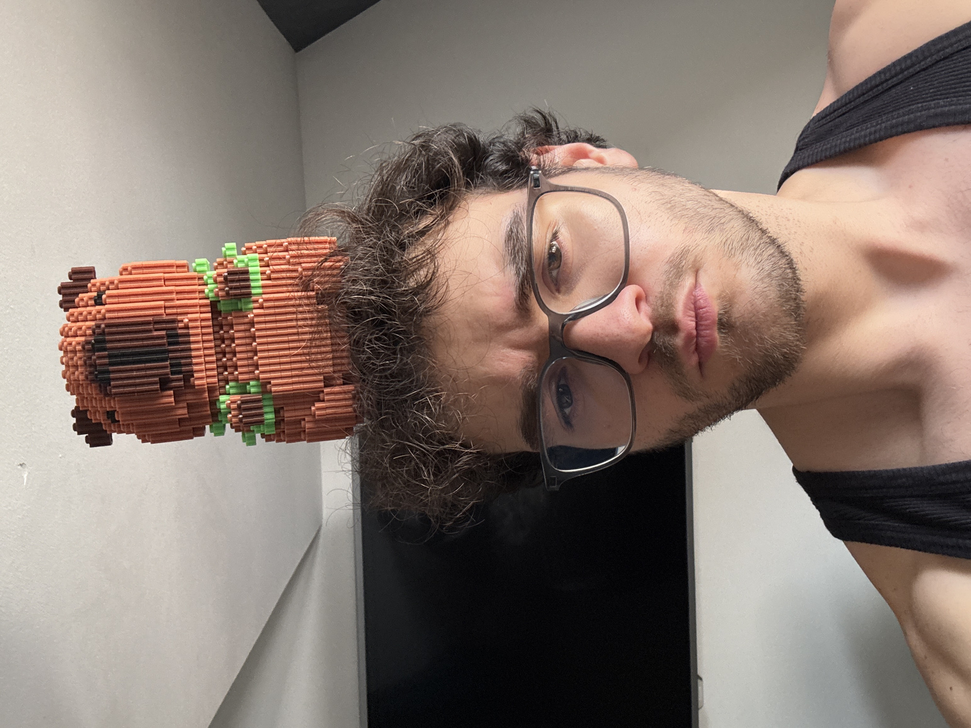 The media file presents a close-up portrait of a young man, rotated 90 degrees clockwise. His head is positioned in the upper part of the frame, looking directly at the viewer with a neutral or slightly serious expression. He has dark, curly hair and light stubble on his face, and is wearing dark-framed eyeglasses. A black ribbed sleeveless top is visible on his torso, which occupies the lower right portion of the image.
Prominently featured to the left of the man's head is a large, intricate pixel art sculpture. This figure, made from numerous small, rectangular blocks, primarily in shades of brown and black with some light green accents, depicts a cartoonish dog-like character. It features distinct ears, a snout, and a green collar, reminiscent of characters like Scooby-Doo.
The background is simple, consisting of light grey walls. A dark, possibly black, rectangular object, perhaps a screen or piece of furniture, is visible in the lower right background behind the man's shoulder. The lighting is even and soft, suggesting an indoor setting. No discernible text is visible.
