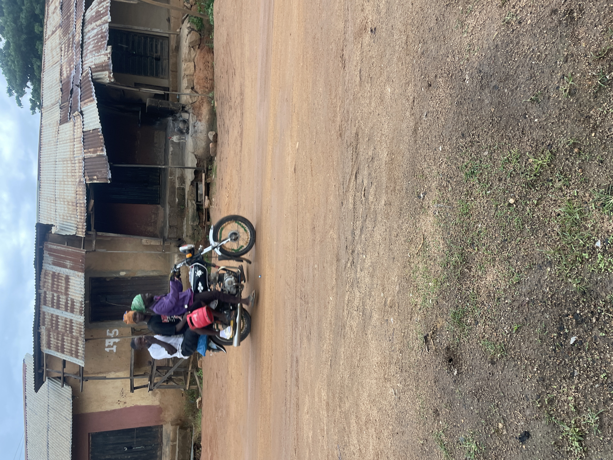 The image shows three young men riding on a motorcycle on a dirt road in Jos, Nigeria. The man in the front is wearing a purple jacket and a green headscarf. The middle rider is wearing a black shirt and an orange headscarf. The man in the back is wearing a white t-shirt. A red fuel container is strapped to the back of the motorcycle. In the background, there are simple buildings with corrugated iron roofs. The sky is overcast. The overall scene depicts a typical street view in Jos, Nigeria.