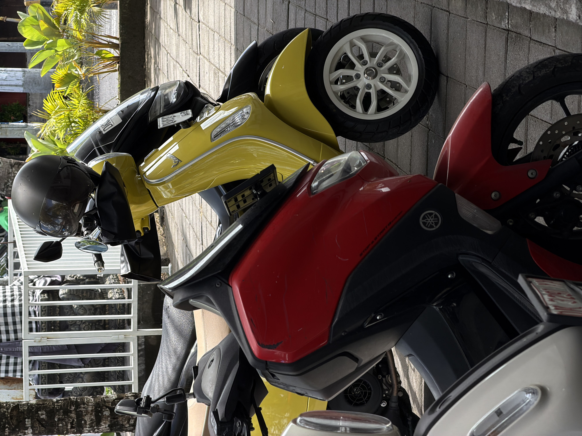 The image depicts a collection of motorcycles parked on a paved surface. The foreground features a red Yamaha scooter, partially visible, with a scuffed front panel. Adjacent to it is a yellow three-wheeled scooter with a black windscreen and a white front wheel. A helmet rests on the handlebar. In the background, a white metal fence is visible, partially obscuring a patterned cloth. Lush green vegetation grows near a building. The location is North Kuta, Indonesia.
