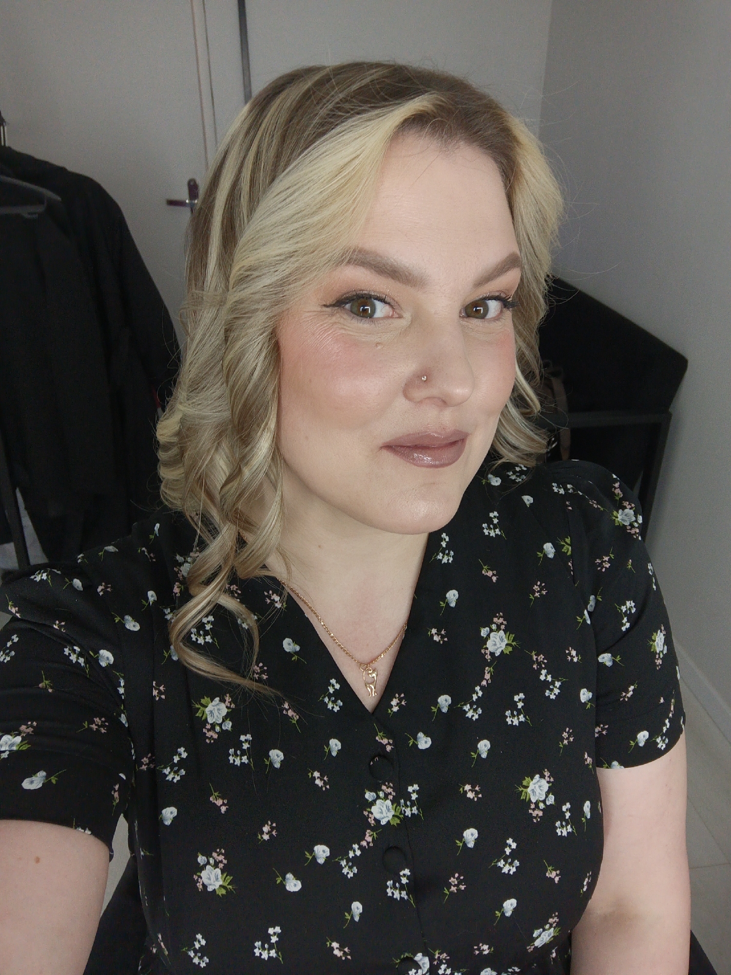 A medium close-up portrait captures a woman from the chest up, looking directly at the camera with a gentle, inviting smile. She appears to be in her late 20s to early 30s. Her hair is a mix of blonde and light brown highlights, styled in soft, defined curls that frame her face. She has brown eyes and is wearing natural-looking makeup, including soft eyeshadow, subtle eyeliner, blush, and a glossy, nude-pink lip color. A small, silver-toned stud is visible in her right nostril piercing.
She is dressed in a short-sleeved, V-neck black top or dress, which is covered in a delicate pattern of small white, light blue, and pink flowers. Small black buttons run down the front of the garment. Around her neck, she wears a thin gold-colored chain with a small, intricate pendant that appears to be a stylized figure or animal.
The background is simple and uncluttered, primarily a light grey wall. To the left, a dark piece of clothing hangs on a hanger, suggesting an indoor setting like a bedroom or dressing area. No text is visible in the image. The overall mood conveyed is calm and friendly.