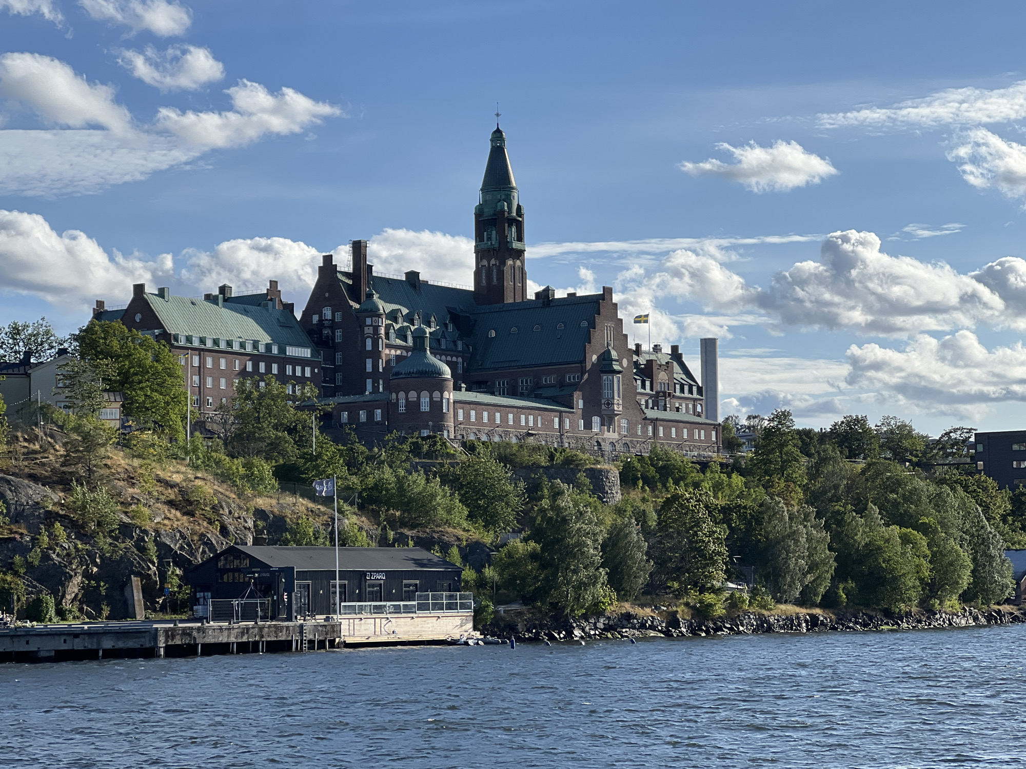 The image shows a large brick building with a green roof and a tall tower, situated on a hill overlooking a body of water. The building appears to be a hotel or a large institution. The hill is covered with trees and rocks. In the foreground, there is a wooden pier with a black building on it, and a flag is flying from a pole. The water is blue and calm, with some ripples on the surface. The sky is blue with white clouds. The building in the background is believed to be the Grand Hôtel Saltsjöbaden in Nacka, Sweden. There are no people clearly visible in the image, and no specific actions or interactions are depicted.