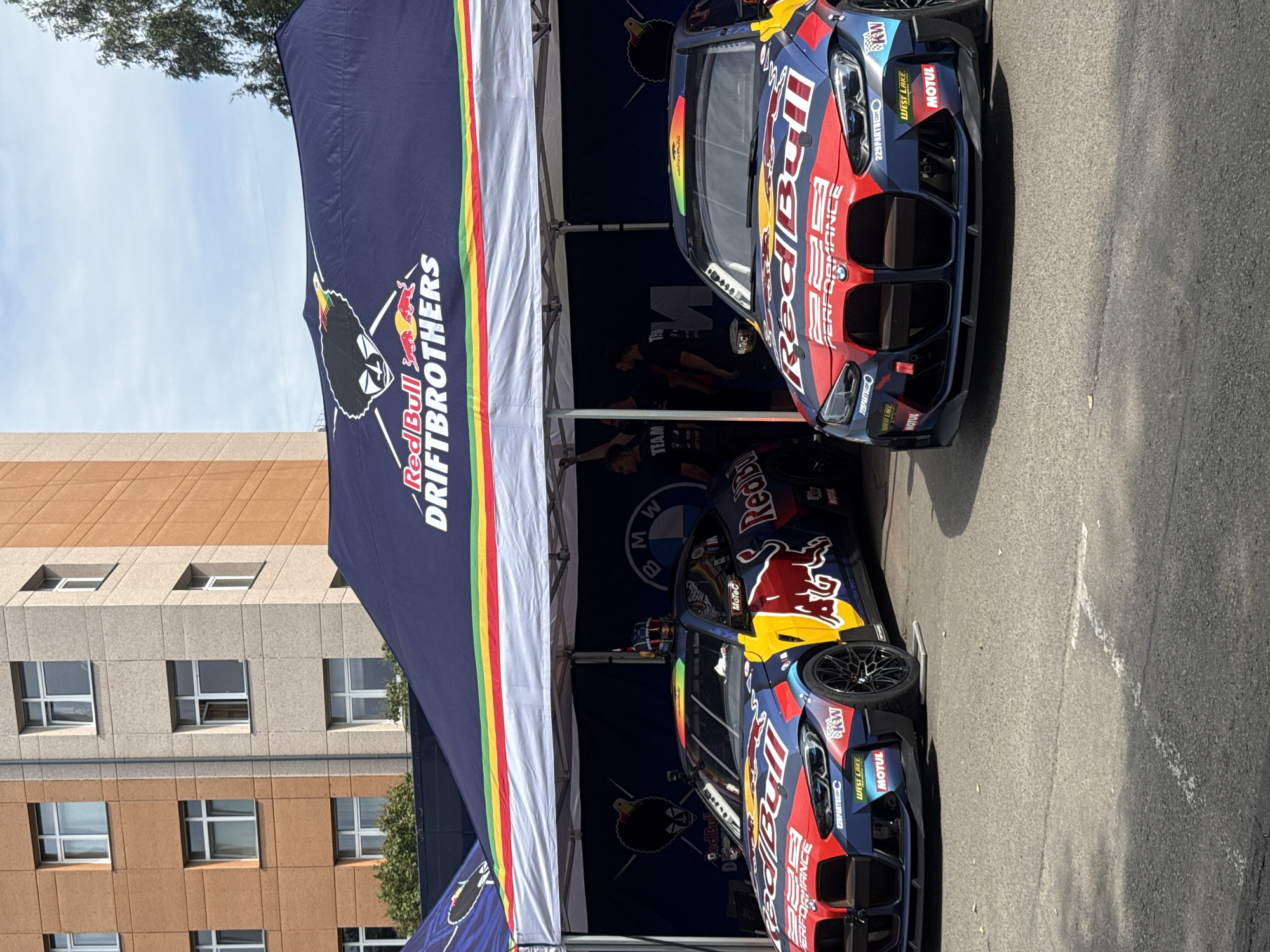 Two BMW M4 racing cars, heavily branded with "Red Bull Driftbrothers" livery and sponsors like Motul and KW, are parked under a large blue tent. The tent features the Red Bull Driftbrothers logo and a rainbow-colored stripe along its bottom edge. Behind the tent, a multi-story building with tan and light gray facade and numerous windows is visible. Several people, seemingly part of the racing team, are standing or walking under the tent. The scene is set on an asphalt surface, possibly a pit lane or parking area, with some white markings on the ground. The sky above is partly cloudy. The specific location in Riga, Latvia is unknown.
