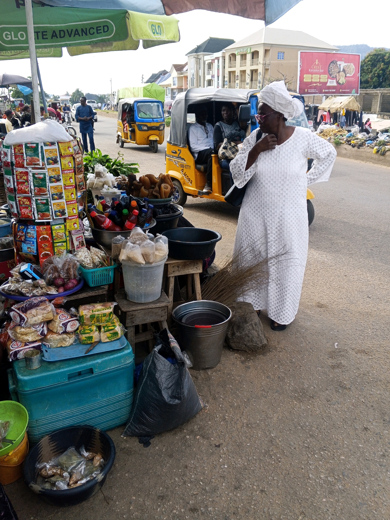 This image captures a lively street scene in Lokoja, Nigeria. In the foreground, a woman in a white, patterned dress and headscarf stands near a street vendor's stall. She is looking to her right, with her hand touching her chin, possibly contemplating a purchase or engaging in conversation. The vendor's stall is abundant with goods, including loaves of bread, packaged food items with visible text like "TASTY TOM" and "BENNY," bottles of drinks, and various other bagged and loose produce. A large blue cooler and several buckets are also part of the display.
Behind the stall and the woman, a yellow and blue auto-rickshaw, known locally as a "keke," is parked. Two men are seated inside, and a third person is partially visible. In the background, other vehicles, including another keke and a car, are on the road. A billboard for "CREST Karaoke Bar" is prominently displayed. The setting appears to be a busy marketplace or street, with buildings lining the road. The sky is overcast, suggesting a daytime scene, possibly before or after a rain shower, as indicated by the dampness on the ground in some areas and the presence of water in a bucket. The overall atmosphere seems to be one of everyday commerce and activity in a Nigerian urban environment.