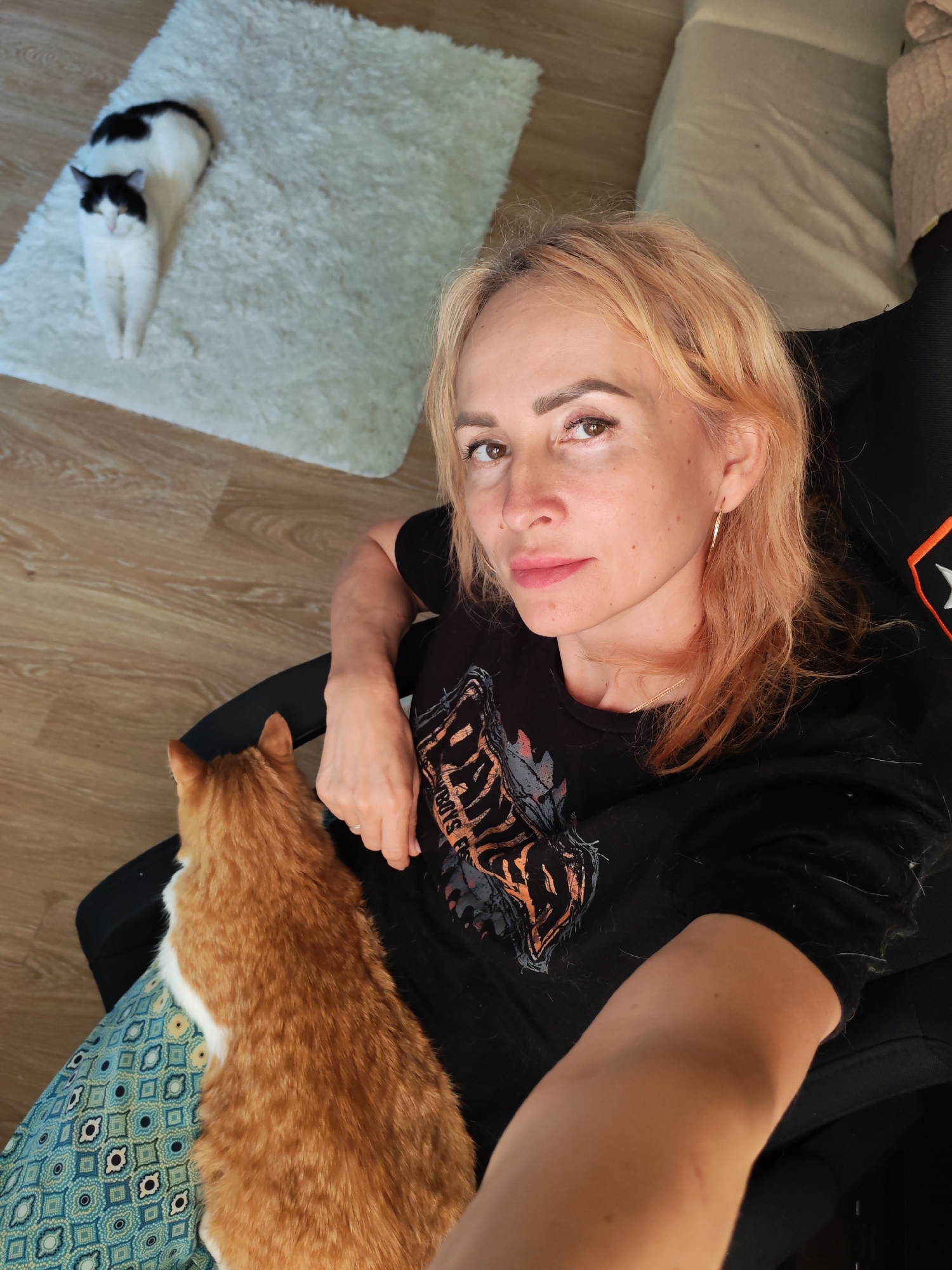 A woman with blonde hair sits in a black chair, holding an orange and white cat on her lap. The cat is resting on a blue and green patterned cushion. To the left of the woman, on a white fluffy rug, a black and white cat is lying down. The floor is made of light-colored wood. The scene is indoors, in a home setting. No specific location in Chaikovsky, Russia is visible or implied by the image content.