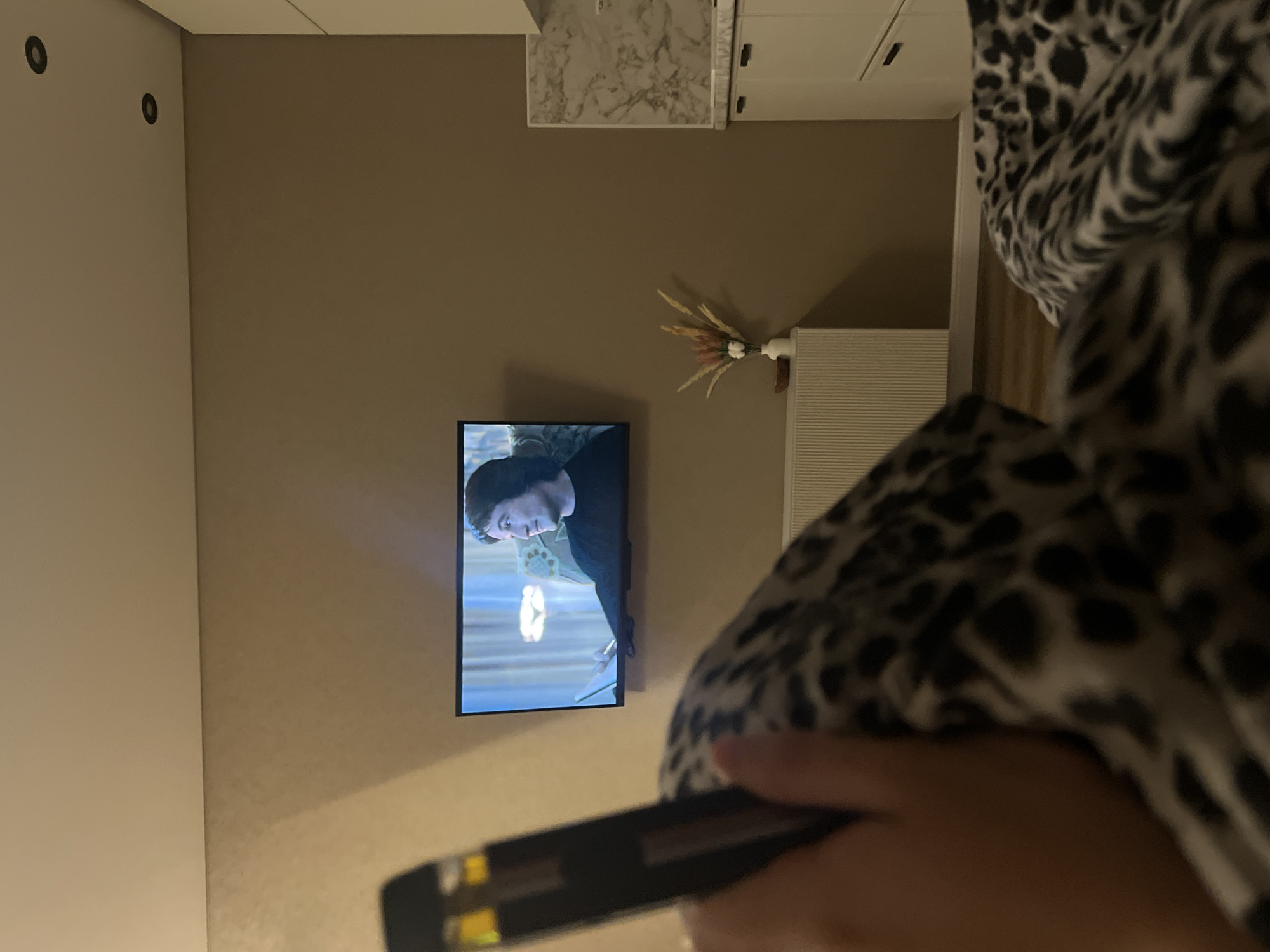 An indoor scene shows a hand holding a black object, possibly a vape pen, in the foreground. To the left, a leopard-print blanket is visible. In the middle ground, a flat-screen television mounted on a tan wall displays a man watching something. Below the television, a white radiator is partially obscured by the blanket. To the right, a kitchen counter with white cabinets and a marble backsplash is visible. The scene appears to be a living room or bedroom area adjacent to a kitchen, likely in an apartment. The image does not provide specific details to pinpoint a location within Krasnodar, Russia.