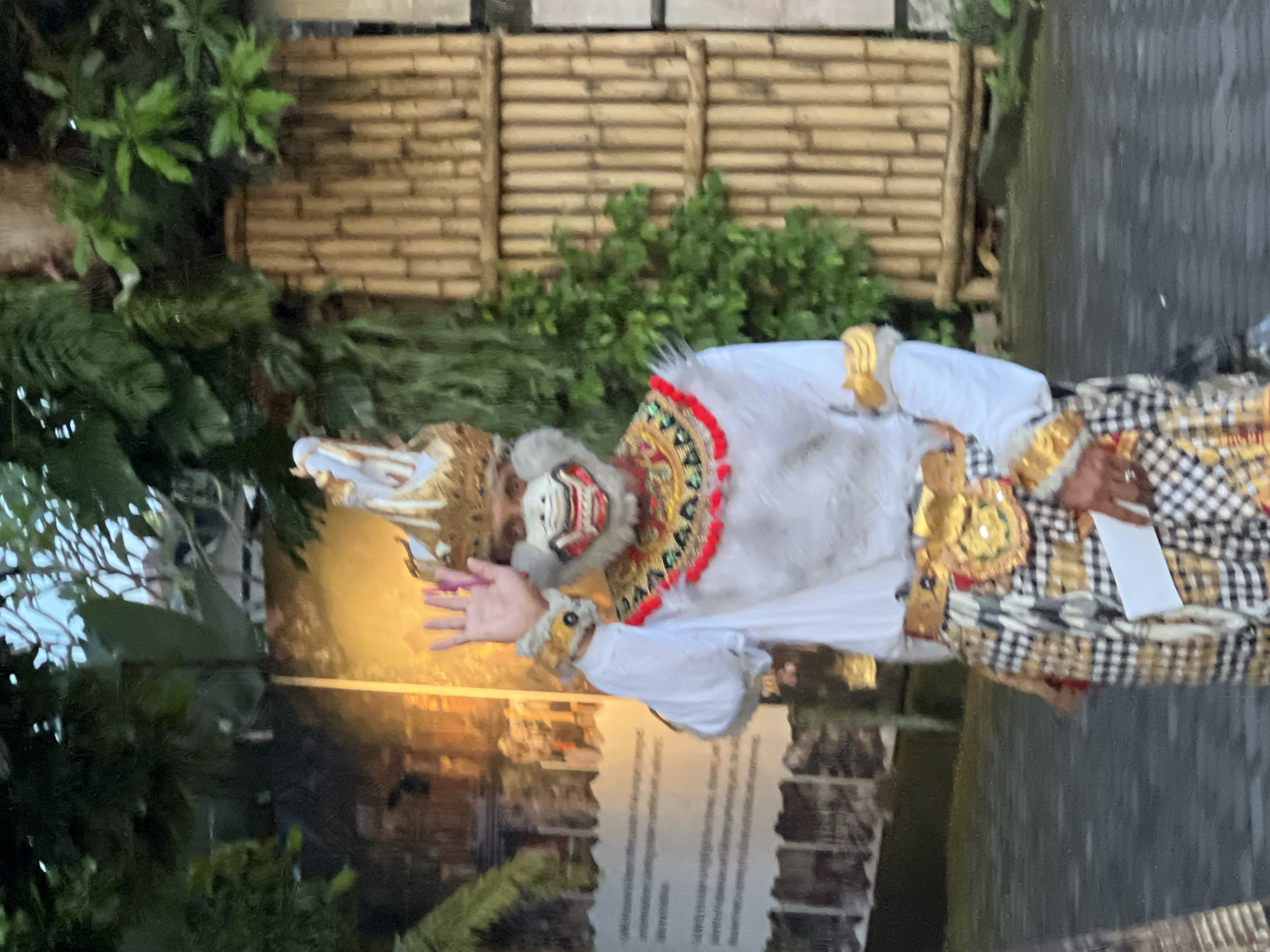 A photograph of a person in Ubud District, Indonesia, wearing a costume consisting of a headdress, white shirt, and black-and-white checkered sarong, and a mask with red and gold accents. The person is holding out one hand. The background includes greenery and a sign with printed text.