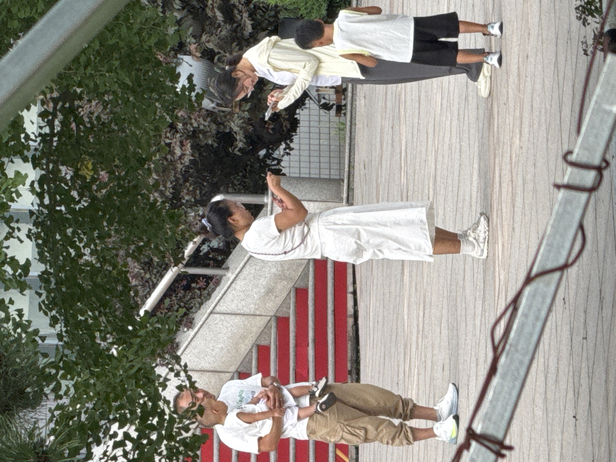 In Zhongtun, China, a group of four people stands on a paved outdoor area with trees and a building in the background. On the left, a man wearing a white t-shirt and khaki cargo pants holds a young child dressed in a white onesie. The man is looking down at the child. Next to him, a woman in a white dress stands with her head bowed, looking at her phone. To the right, a woman in a light-colored hooded jacket and dark pants also looks down at her phone. A young boy in a white t-shirt and black shorts stands facing away from the viewer. Red stairs are visible behind the man and child. A metal structure with red rope is in the foreground.