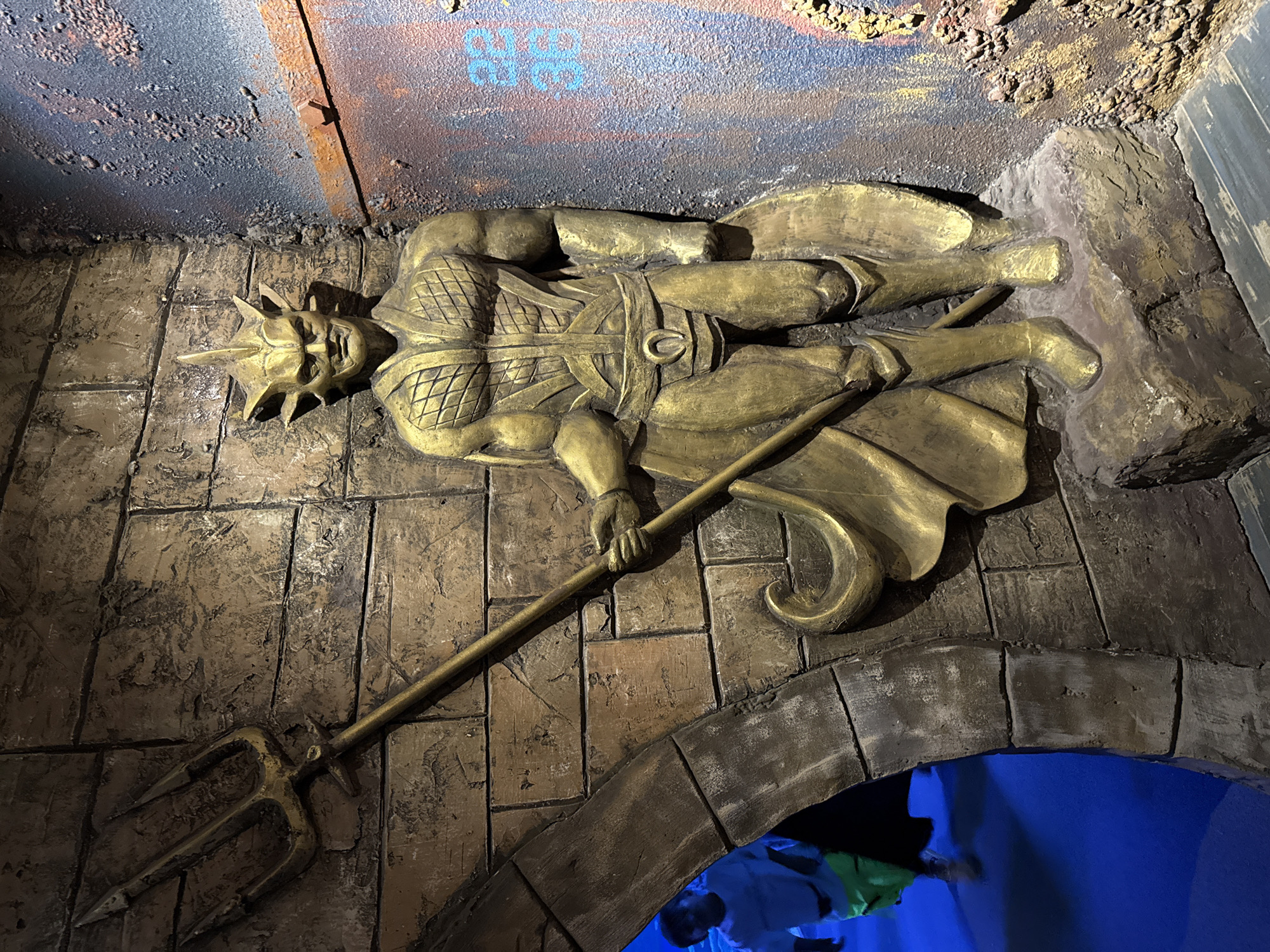 A gold-colored statue of a muscular, humanoid figure with a trident stands against a textured brick wall. The figure has a distinctive helmet with sharp protrusions on the sides and a central crest. Its chest is sculpted with a diamond pattern, and it wears a belt with a horseshoe-shaped buckle. The statue is positioned in a dimly lit environment with a blue floor visible through an arched opening to the left, where a blurred person in a white shirt and green shorts is walking. To the right of the statue, the wall transitions to a rough, textured surface with brown and blue paint splatters, and some rusted metal elements. The overall scene appears to be an indoor decorative installation, possibly part of an amusement park or themed attraction. The location is not specified within the image.