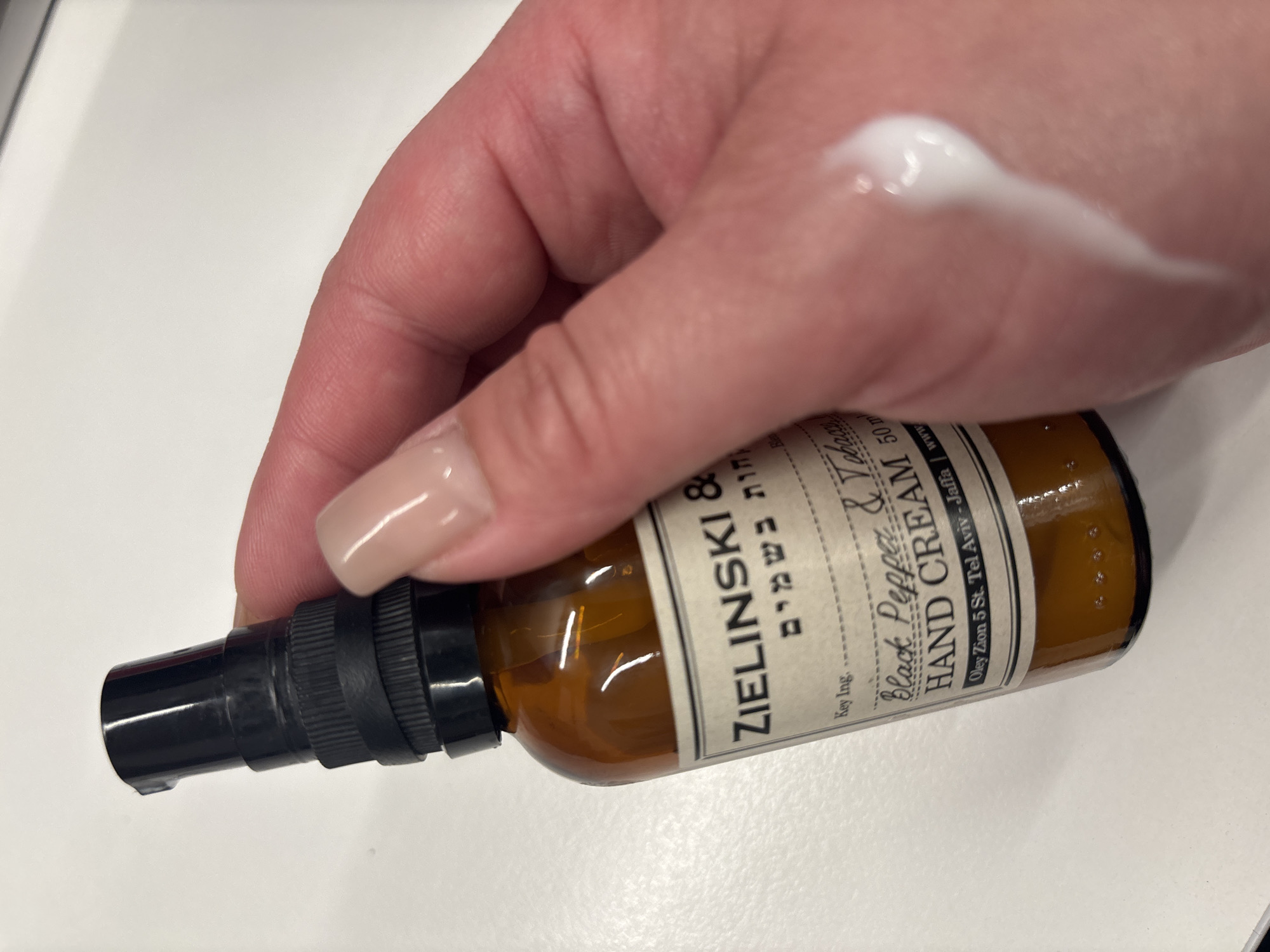 A hand with light skin and neutral-toned, glossy fingernails holds a small, amber glass bottle with a black pump dispenser. The bottle has a paper label with text in English and Hebrew. The English text reads "ZIELINSKI &", "Black Pepper & Tobacco", "HAND CREAM", and "50 ml". Below this is "Oley Zion 5 St. Tel Aviv - Jaff". A dollop of white hand cream is smeared on the back of the hand, near the knuckles. The background is a plain white surface, likely a desk or table. The scene is indoors, under artificial light. The location of Birobidzhan, Russia is not depicted or implied by any visual cues in the image.
