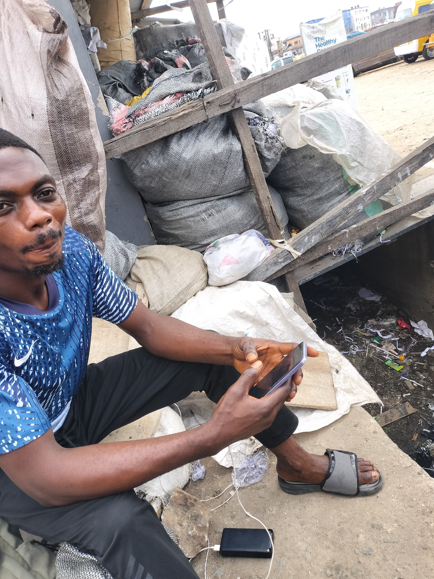The image depicts a man with dark skin and short black hair, wearing a blue patterned Nike T-shirt and black pants, sitting outdoors while using a smartphone. He is positioned on a concrete surface, with a black power bank nearby, connected to his phone. His right foot is clad in a gray sandal. Behind him, there's a collection of large sacks containing various materials, possibly waste or recyclables, stacked against a rudimentary wooden structure. The background shows a street scene, including a building with a sign that reads "The Healthy". The scene appears to be in an urban setting, potentially in Lagos, Nigeria, given the general appearance of the environment. There is visible trash and debris near the edge of the concrete surface where the man is sitting.