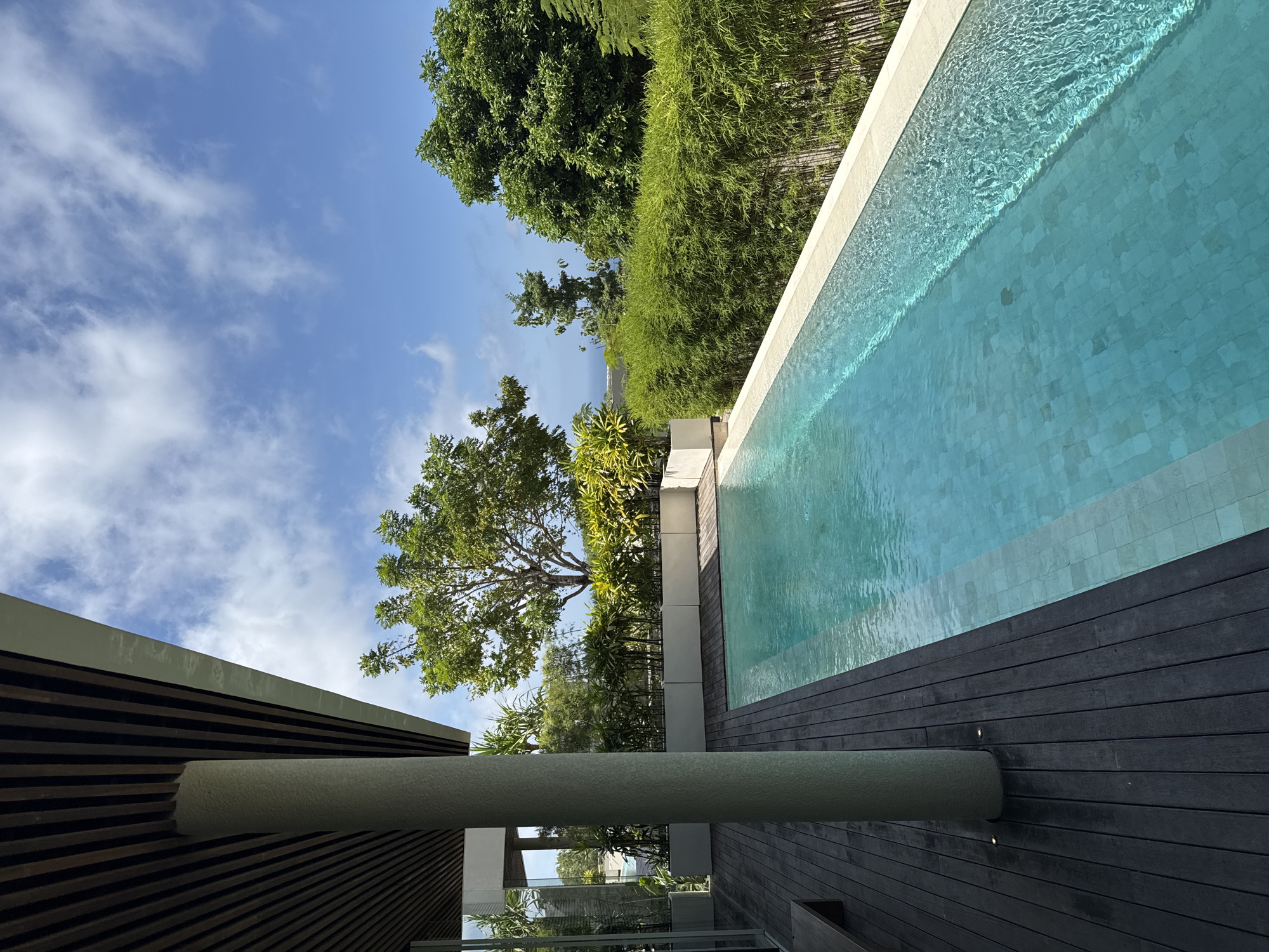The image depicts a pool area adjacent to a building in Kuta Selatan, Indonesia. The pool, filled with water, is lined with light-colored tiles and bordered by a dark wooden deck. The building features horizontal wooden slats and a cylindrical concrete structure attached to its side. Green trees and foliage are visible beyond the pool area, with a blue sky and scattered clouds overhead.