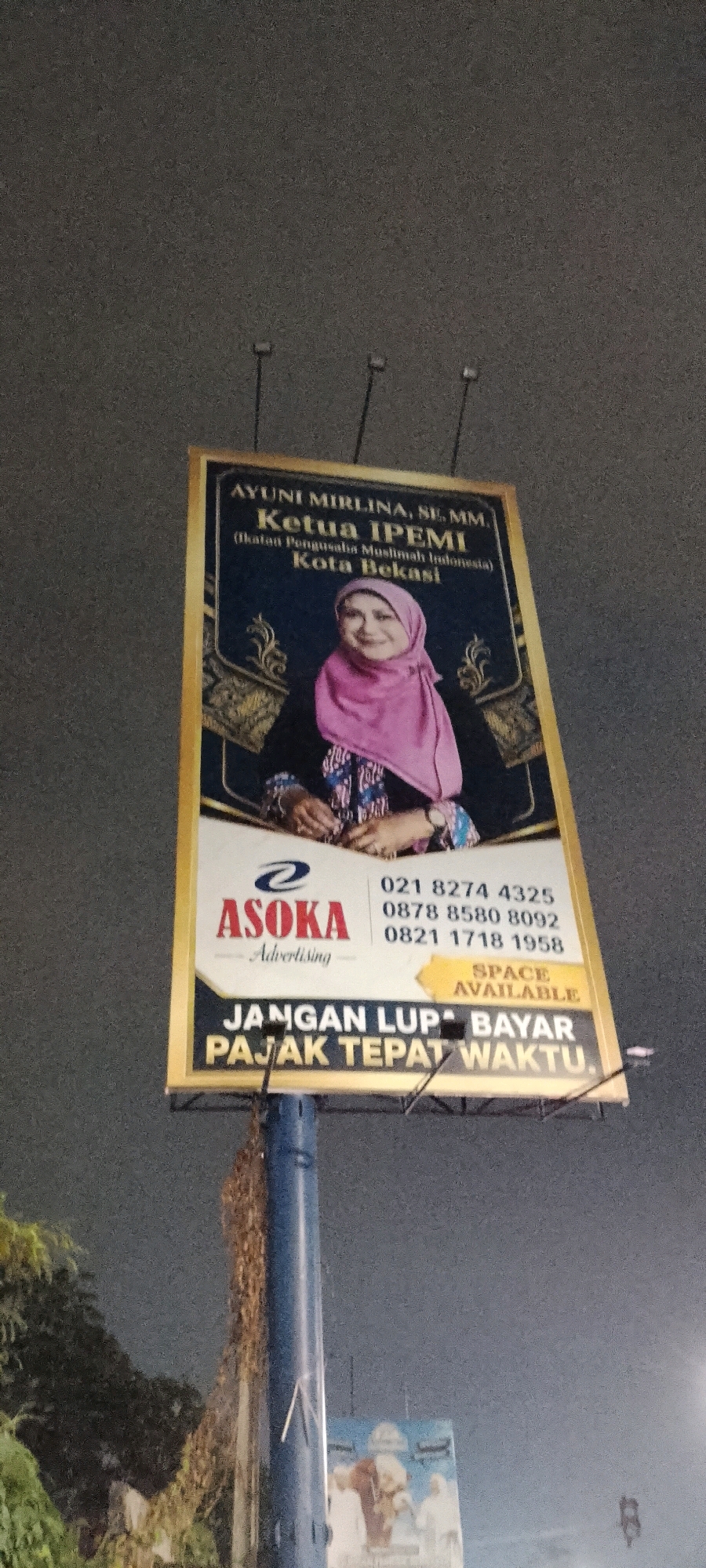 A billboard advertising for Ayuni Mirlina, SE, MM, Ketua IPEMI (Ikatan Pengusaha Muslimah Indonesia) Kota Bekasi is displayed at night. The billboard features a portrait of Ayuni Mirlina, a woman wearing a pink hijab and traditional Indonesian attire. Below her portrait, the advertising company "ASOKA Advertising" is listed with contact phone numbers. The billboard also includes the message "JANGAN LUPA BAYAR PAJAK TEPAT WAKTU," which translates to "DON'T FORGET TO PAY TAXES ON TIME." The billboard is mounted on a blue pole. In the background, a blurry image of another billboard is visible, along with trees and a dark, cloudy sky, suggesting the photo was taken outdoors in an urban environment.